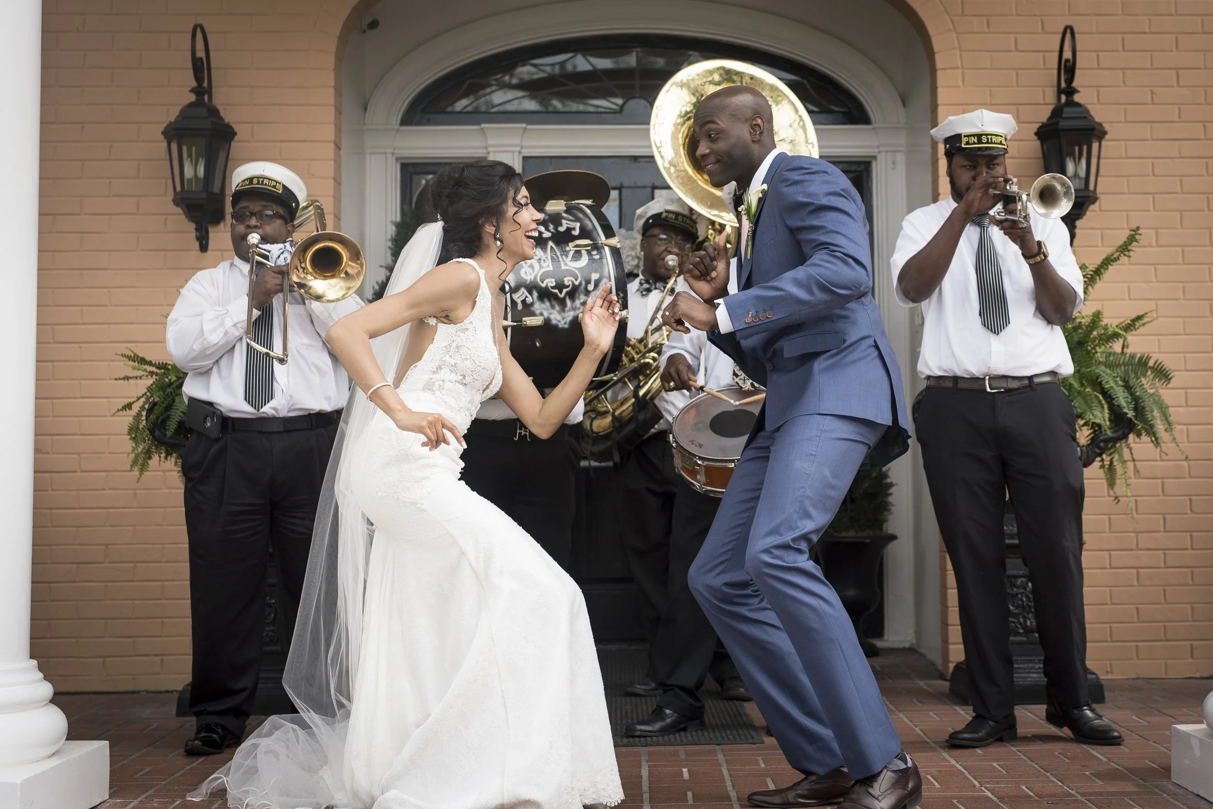 A bride and groom dancing outside in front of a band playing instruments, with a brick building as the background.