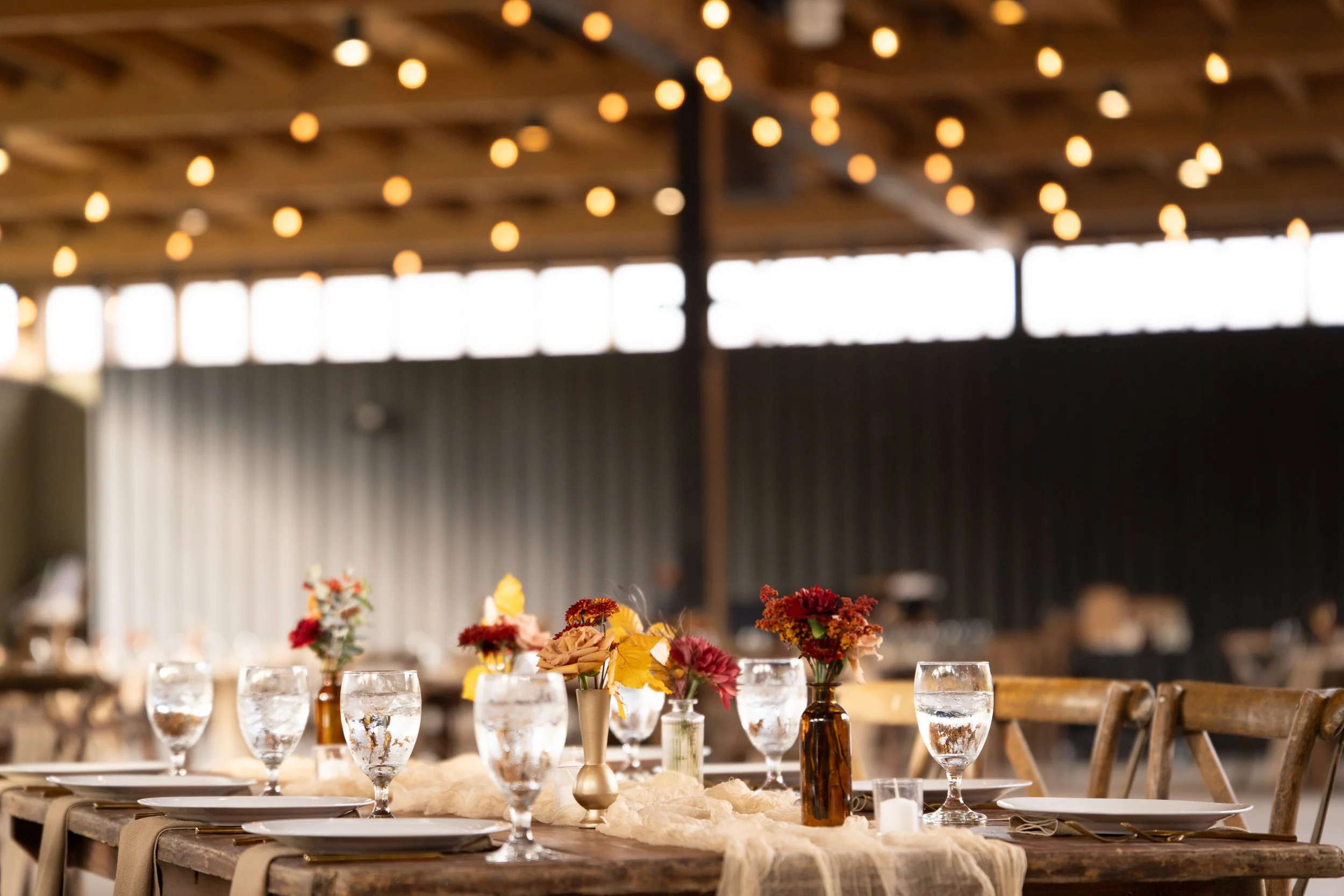 Table decorated with flowers and glassware in a rustic indoor setting with string lights overhead.