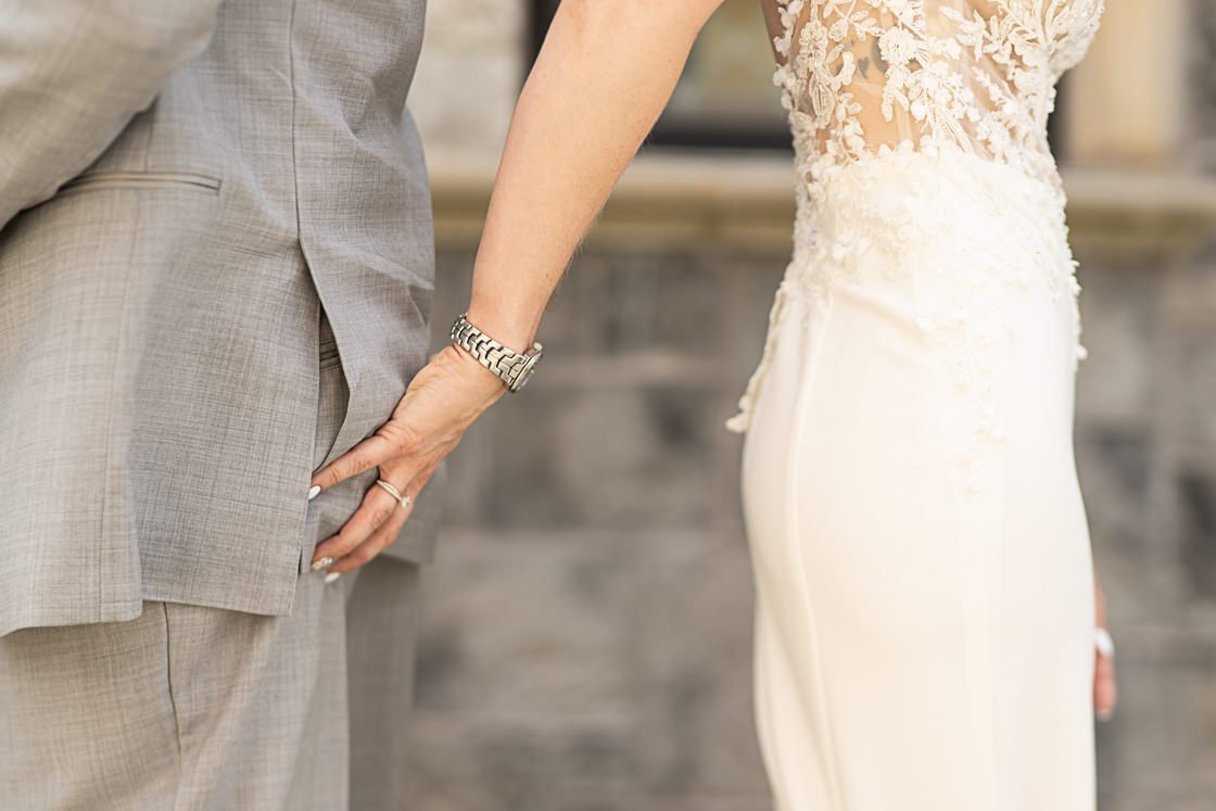 Close-up of a bride and groom holding hands, focusing on their hands and the lower part of their bodies, with a blurred background.