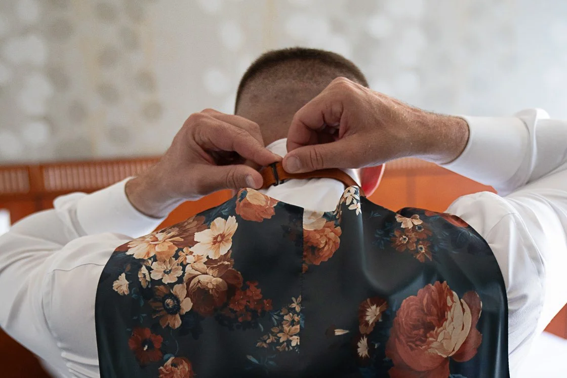 Man adjusting his collar in front of a mirror, viewed from behind, wearing a white shirt and a floral vest with large orange and cream flowers.