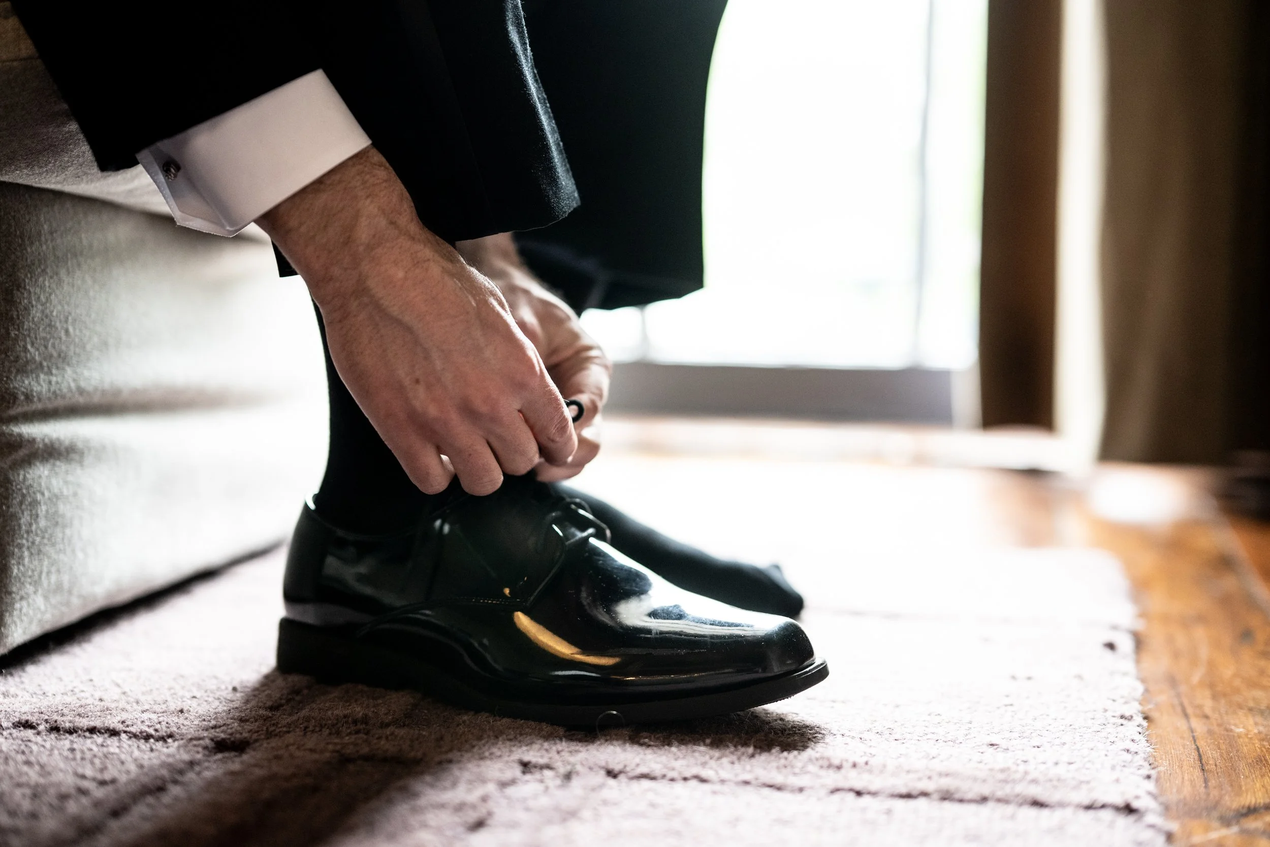 A person in formal attire is tying shoelaces on black shiny dress shoes while standing on a carpeted floor near a window.