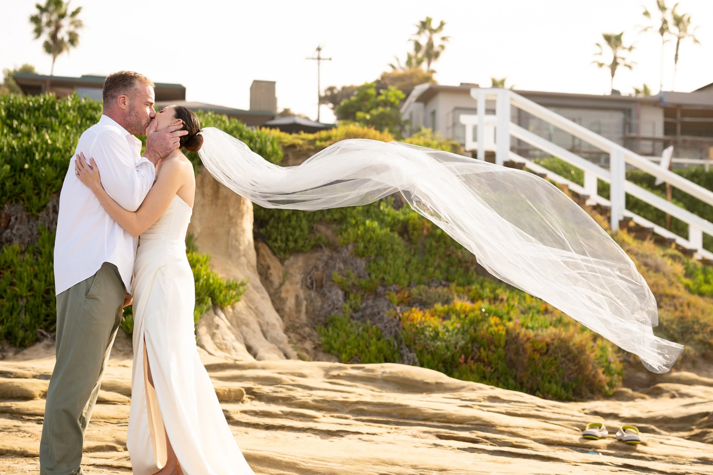 A man and woman sharing a kiss on the beach, dressed in wedding attire, with a flowing veil in the wind. Beach sandals are visible on the sand nearby, and greenery, houses, and palm trees are in the background during sunset.