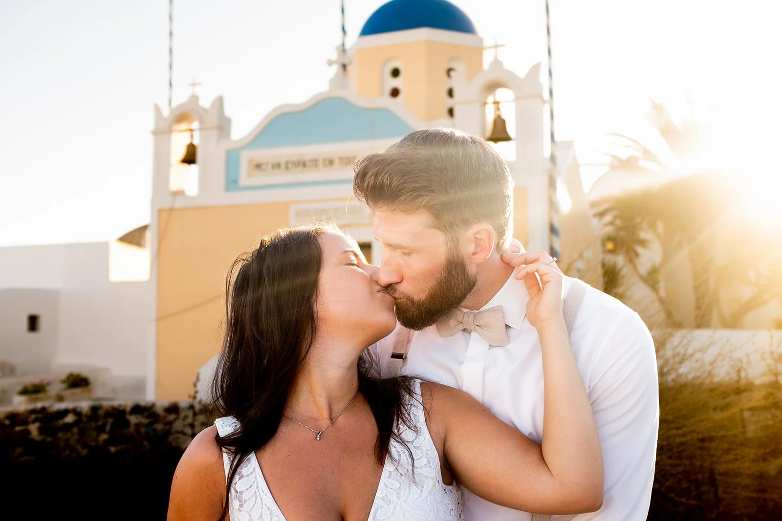 A couple sharing a kiss in front of a church with a blue dome, during sunset.