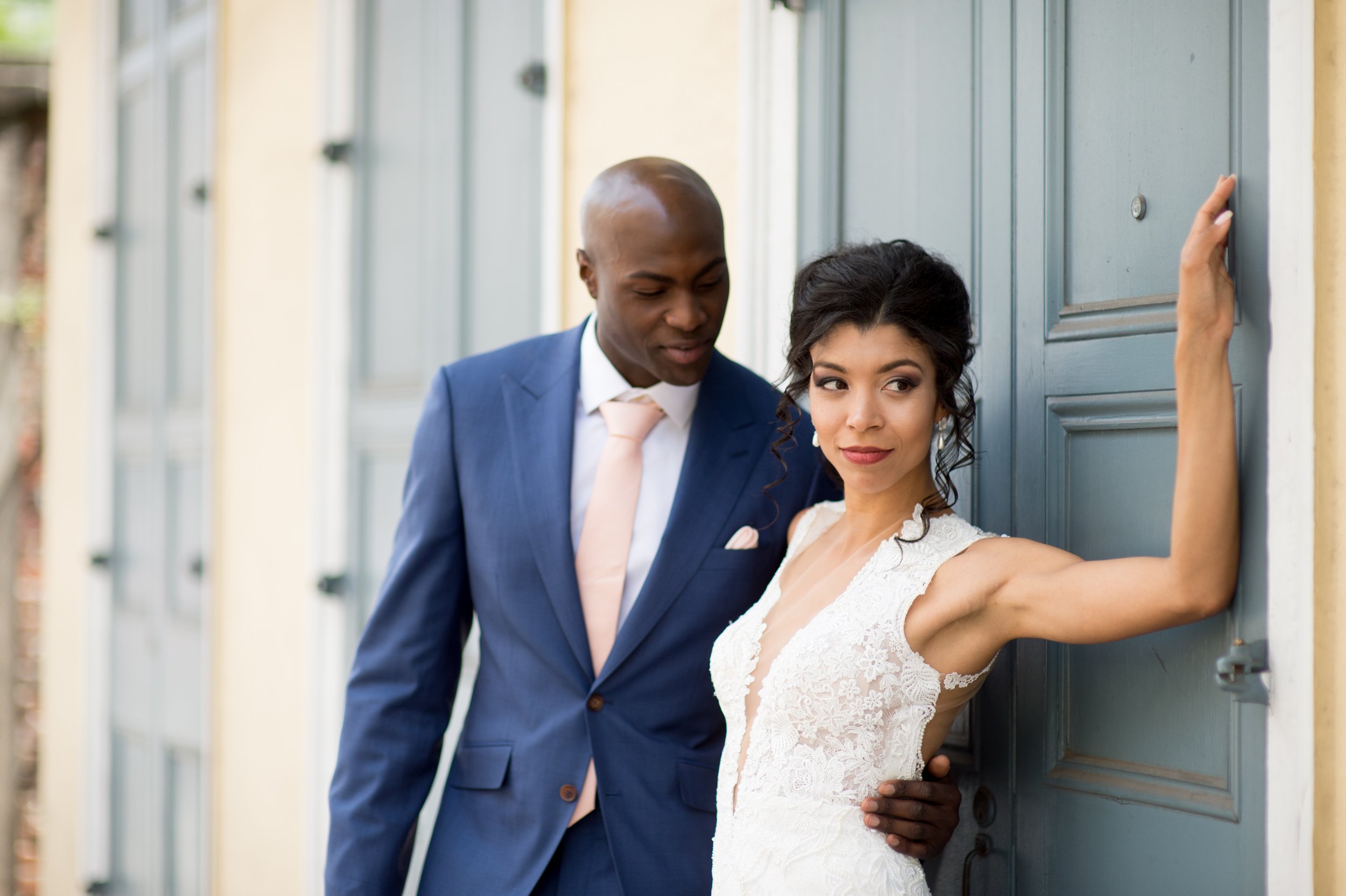 A groom and bride are standing close together near a blue door. The groom is wearing a blue suit with a white shirt and light pink tie, and the bride is wearing a white lace wedding dress. The bride has her left arm resting on the door, and the groom