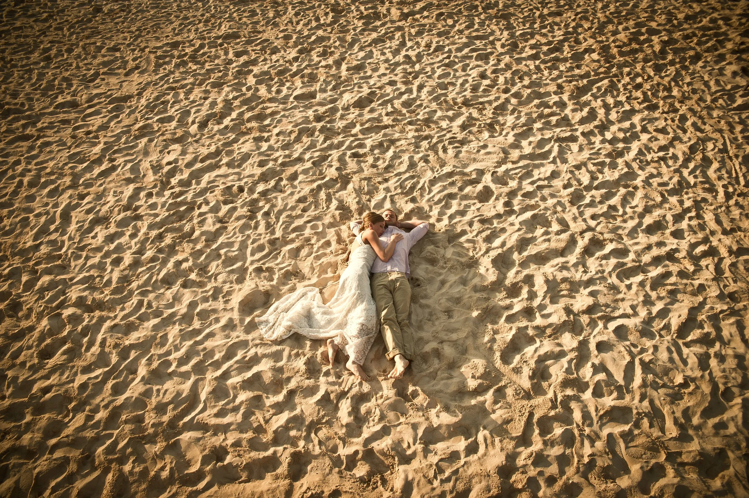 A couple lying on the sand at the beach, with the woman resting her head on the man's shoulder. The sand surrounds them, creating a vast expanse.