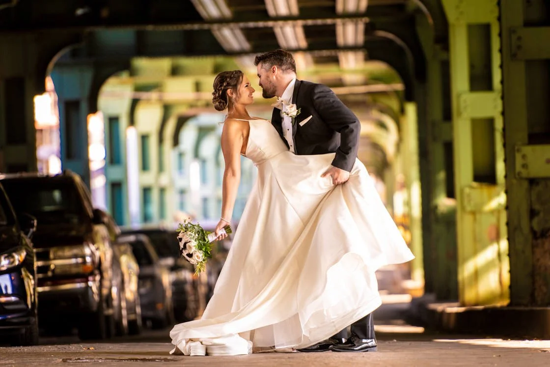A bride and groom sharing a romantic moment under a bridge, the groom lifting the bride's dress, both smiling and looking into each other's eyes.