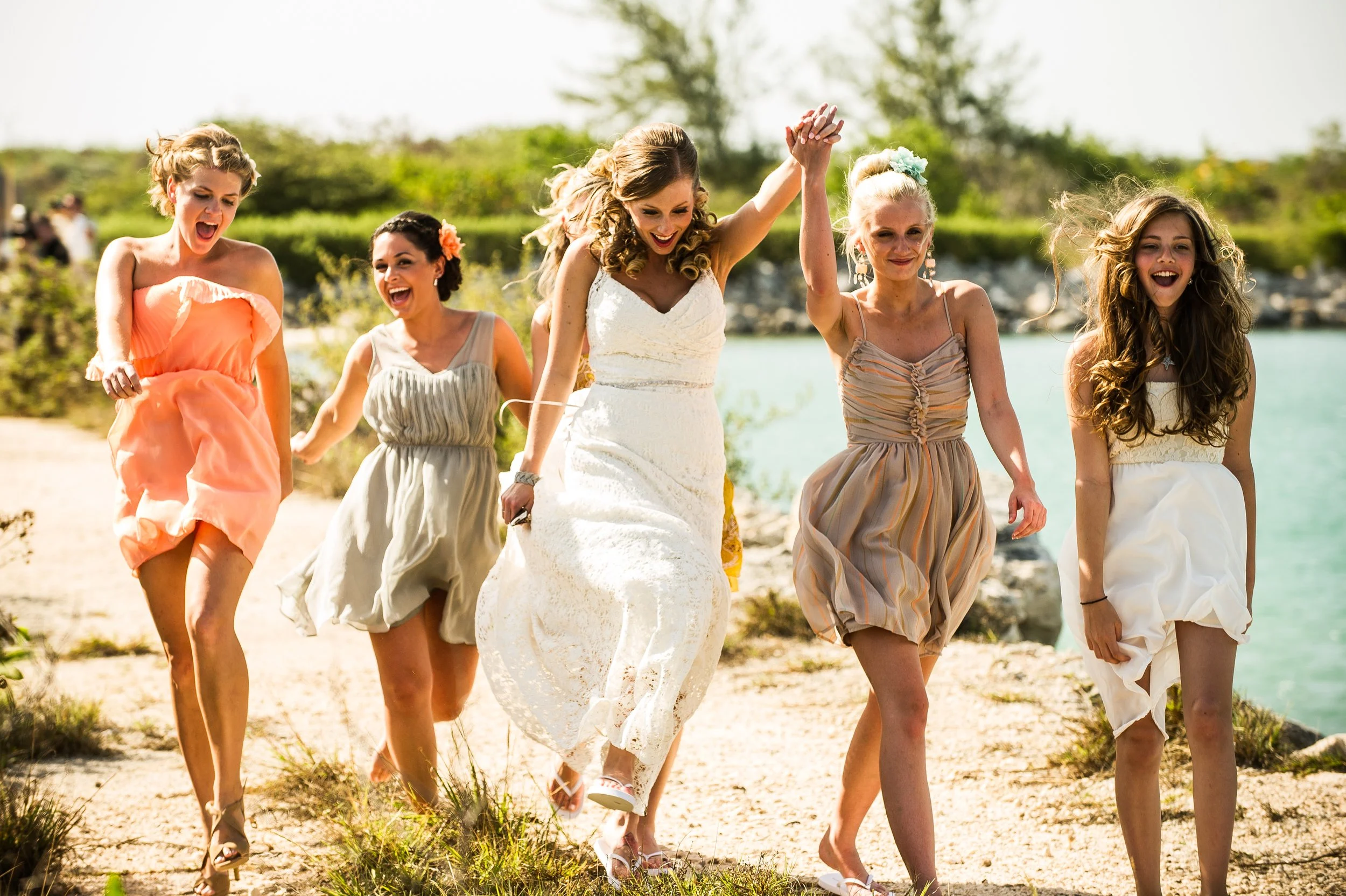 Group of women in dresses walking along a beach with water and trees in the background, celebrating and smiling.