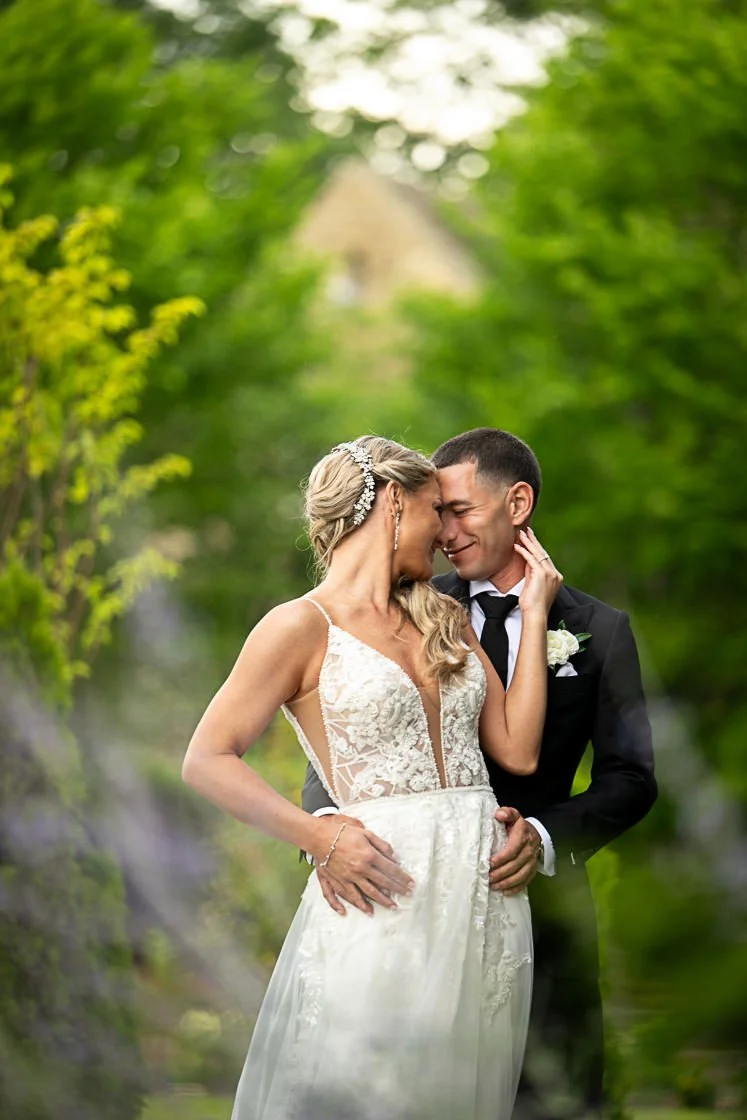 A bride and groom are sharing a tender moment, standing close with their foreheads and noses touching, surrounded by lush green trees in an outdoor setting.