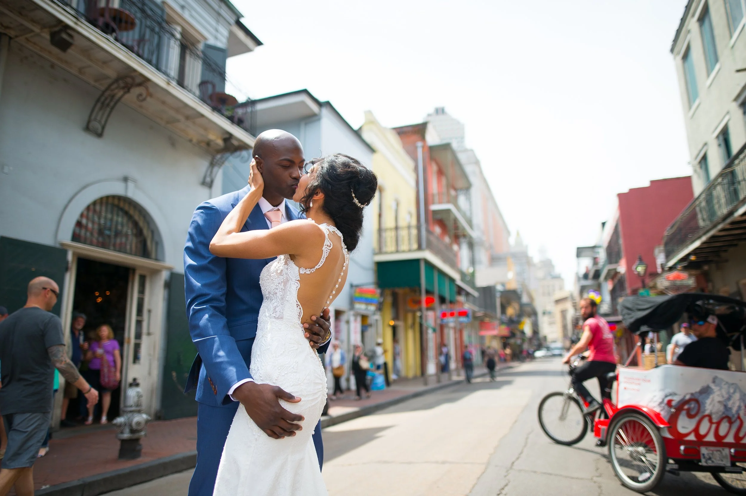 A couple in wedding attire sharing a kiss on a busy city street, with colorful buildings, pedestrians, and bicyclists in the background.