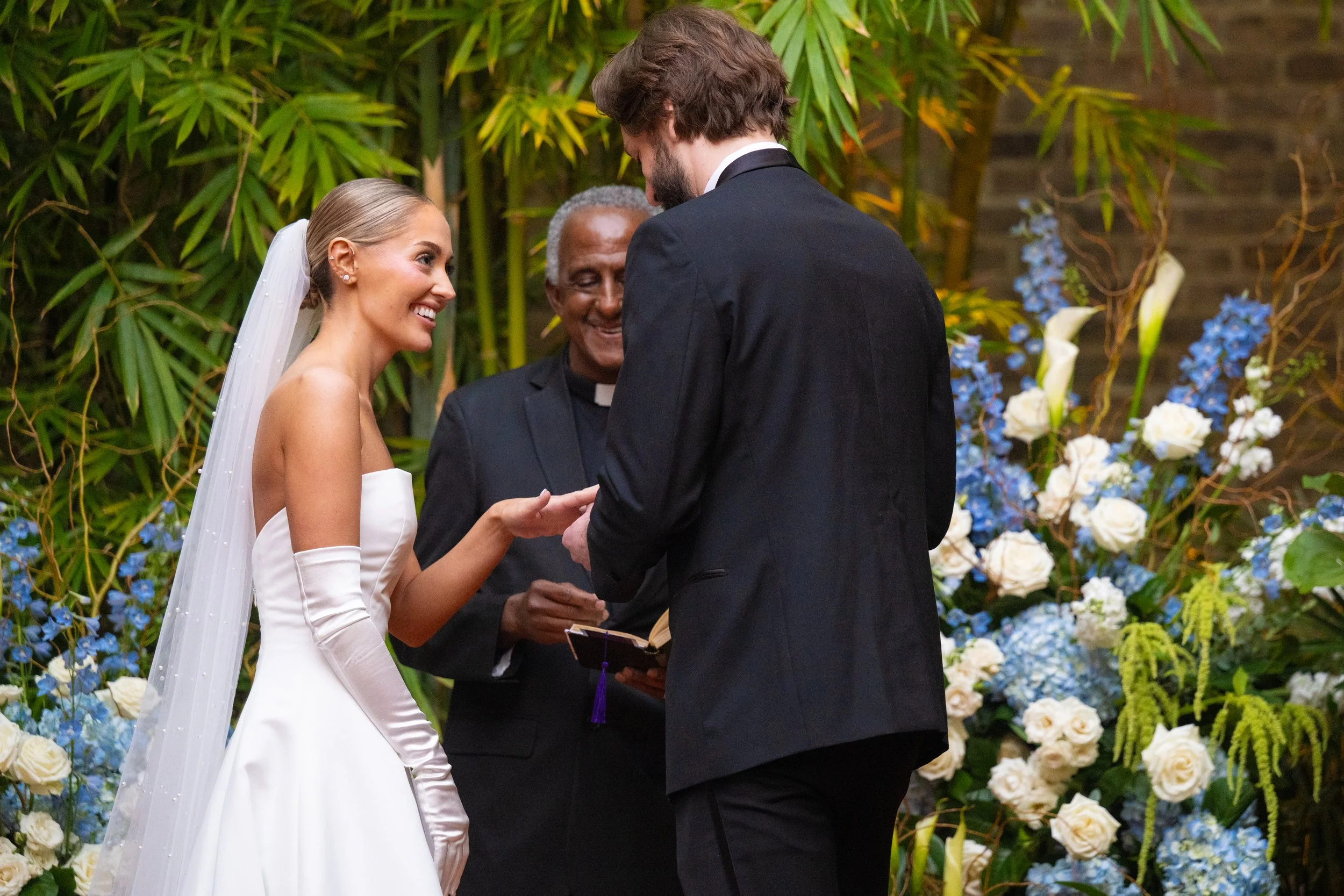A bride and groom exchanging vows during their wedding ceremony, officiated by a clergy member, with a lush backdrop of green leaves and white and blue flowers.