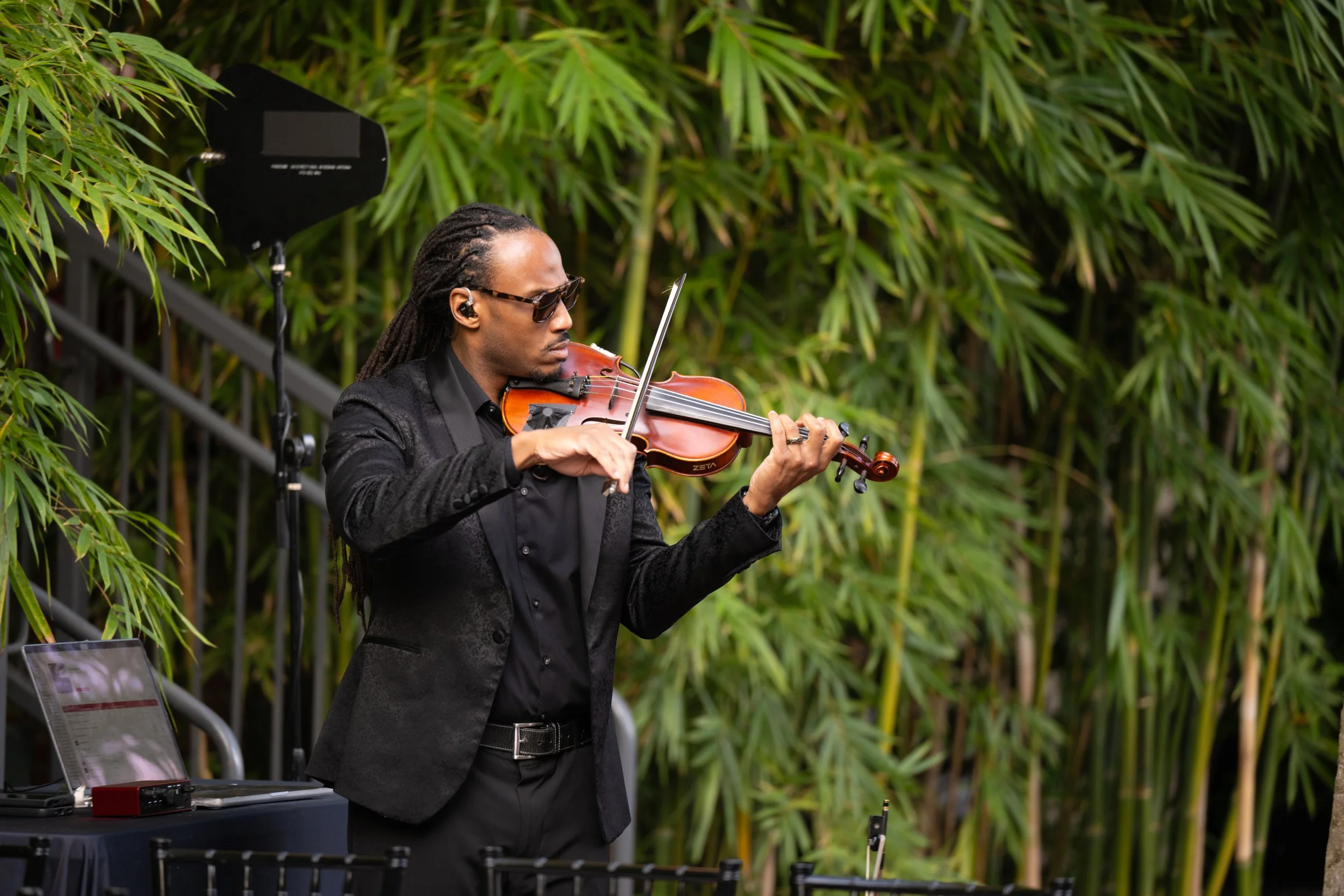 A man playing the violin outdoors surrounded by green bamboo plants.