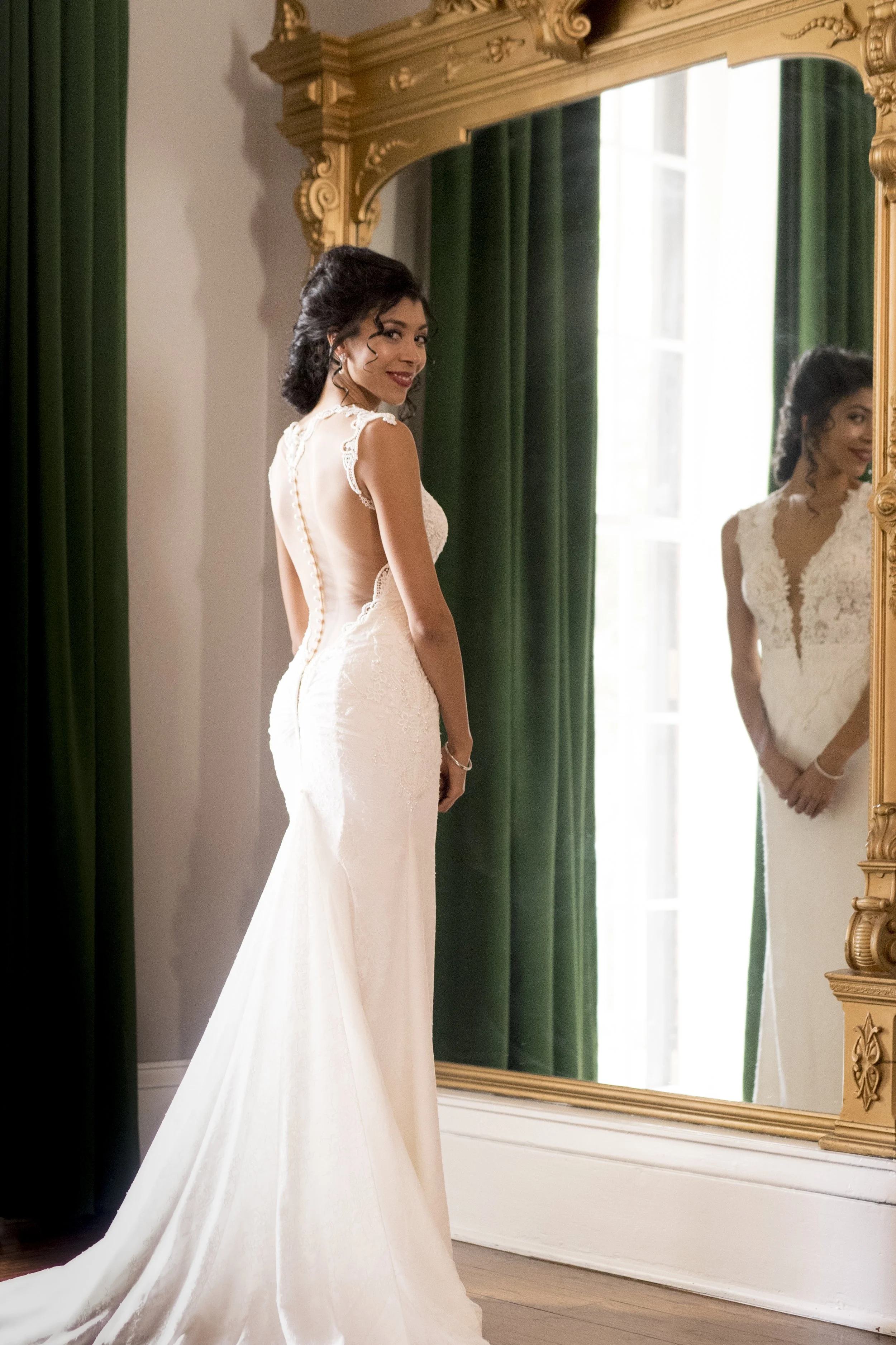 A woman in a white wedding dress standing in front of a large ornate mirror with green curtains behind her. She has dark hair styled in loose curls and is smiling at herself in the mirror.