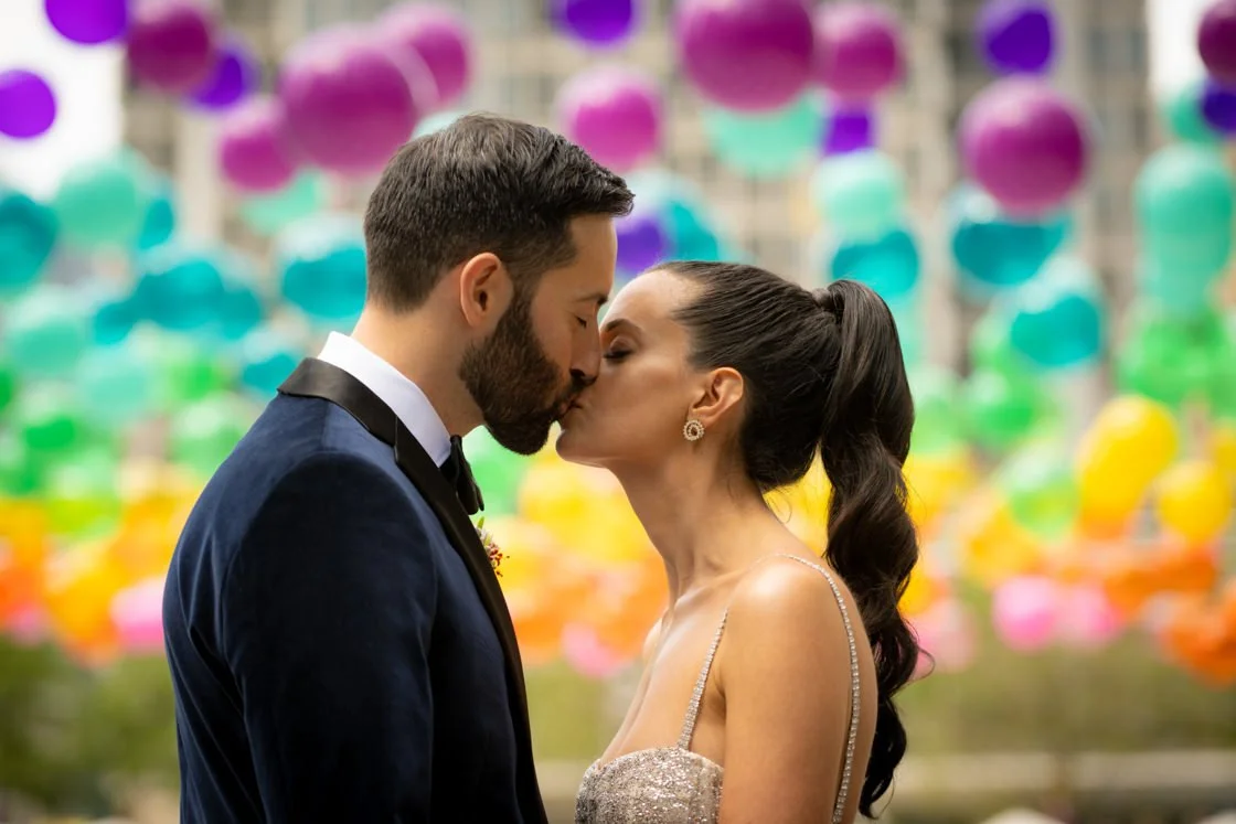 A couple kissing, dressed in wedding attire, in front of colorful balloons.