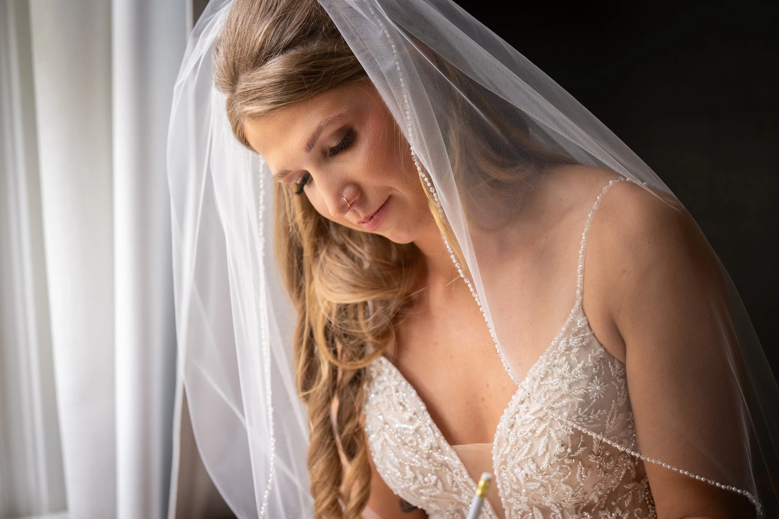 Close-up of a bride with long, wavy blonde hair, wearing a lace wedding dress and a sheer veil, standing near a window with white curtains.