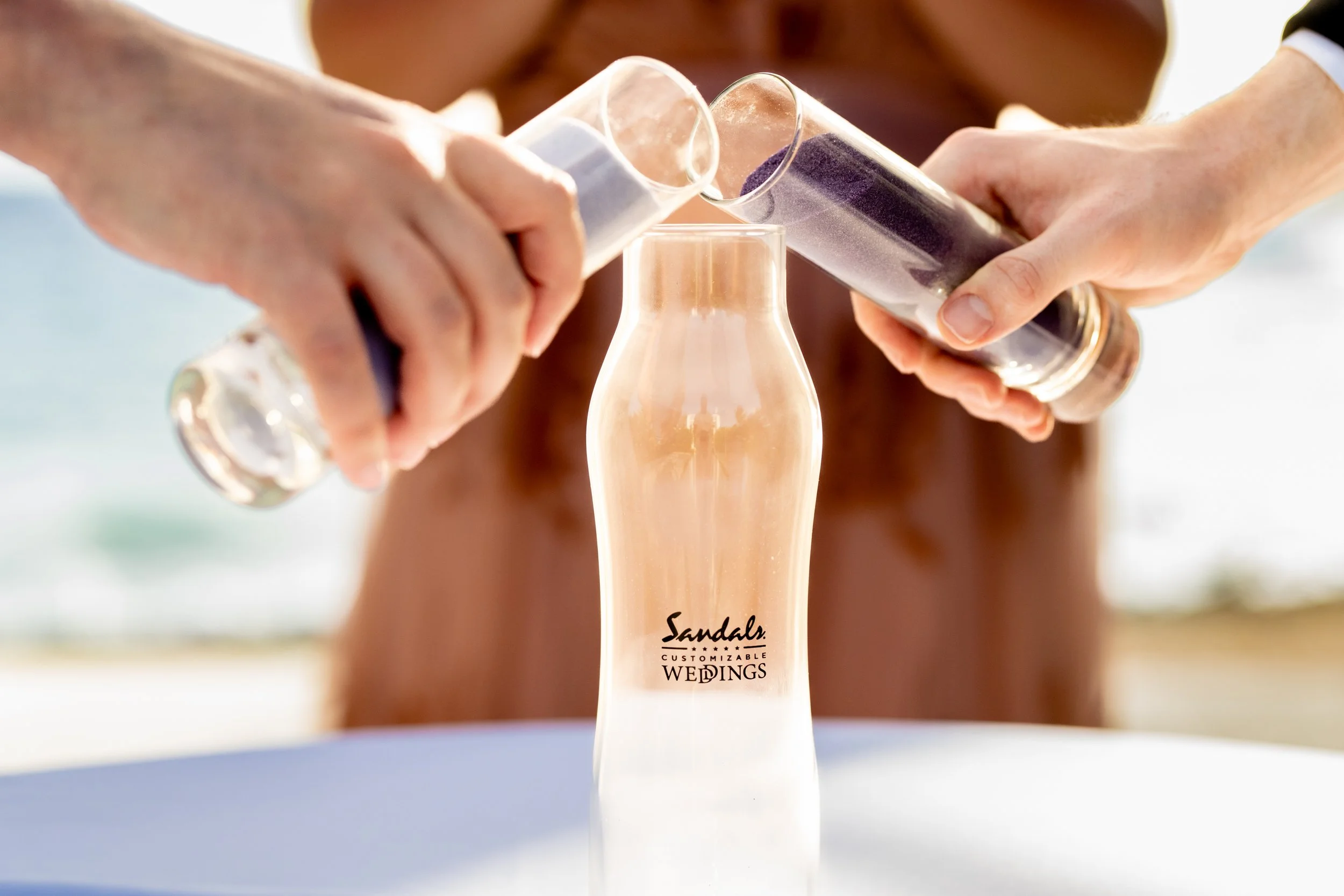 A person pours purple glitter from a test tube onto a champagne flute, with the words 'Sandals Customizable Weddings' on the glass.