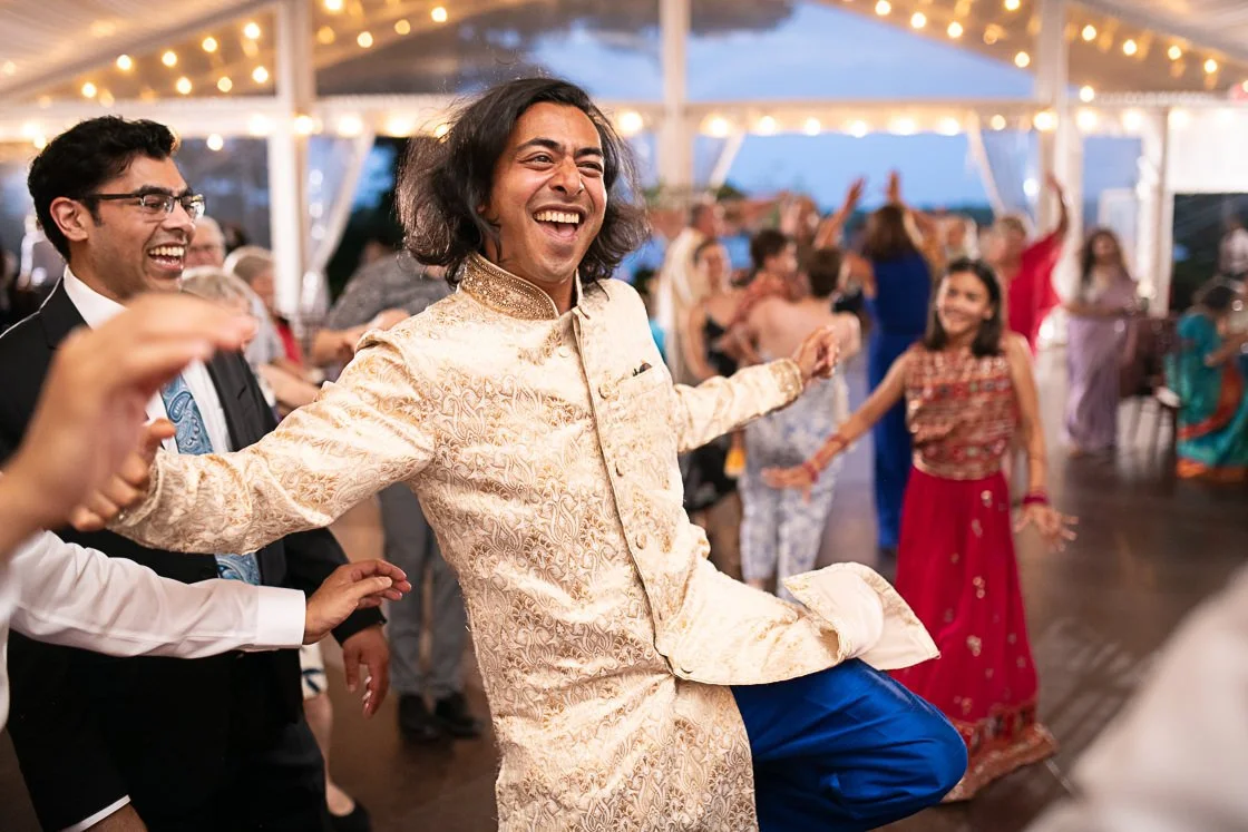 People dancing and celebrating at a festive event under a decorated tent, with string lights overhead, showing joy and happiness.