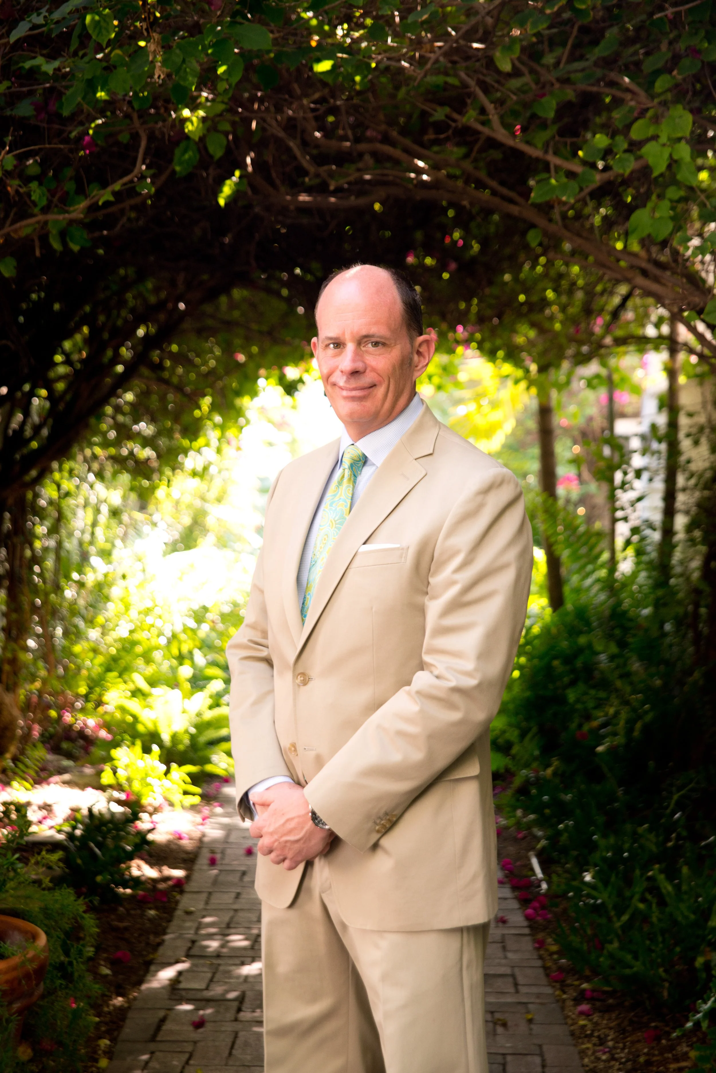 A smiling middle-aged man with a receding hairline in a beige suit and colorful tie, standing on a garden path surrounded by lush greenery and flowering plants.