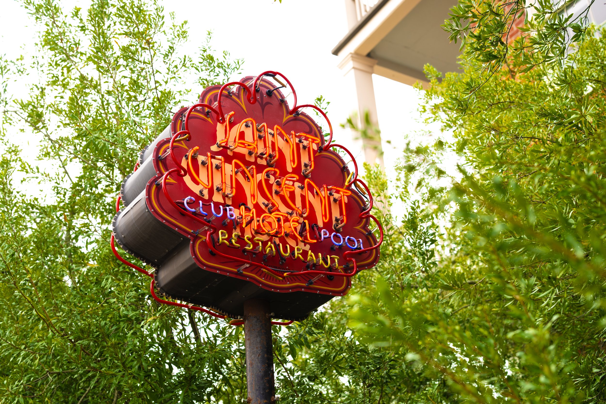 A neon sign for a restaurant called 'Elan' with the words 'Club' and 'Resaurante' visible on it, mounted on a pole surrounded by green trees and foliage in the background.