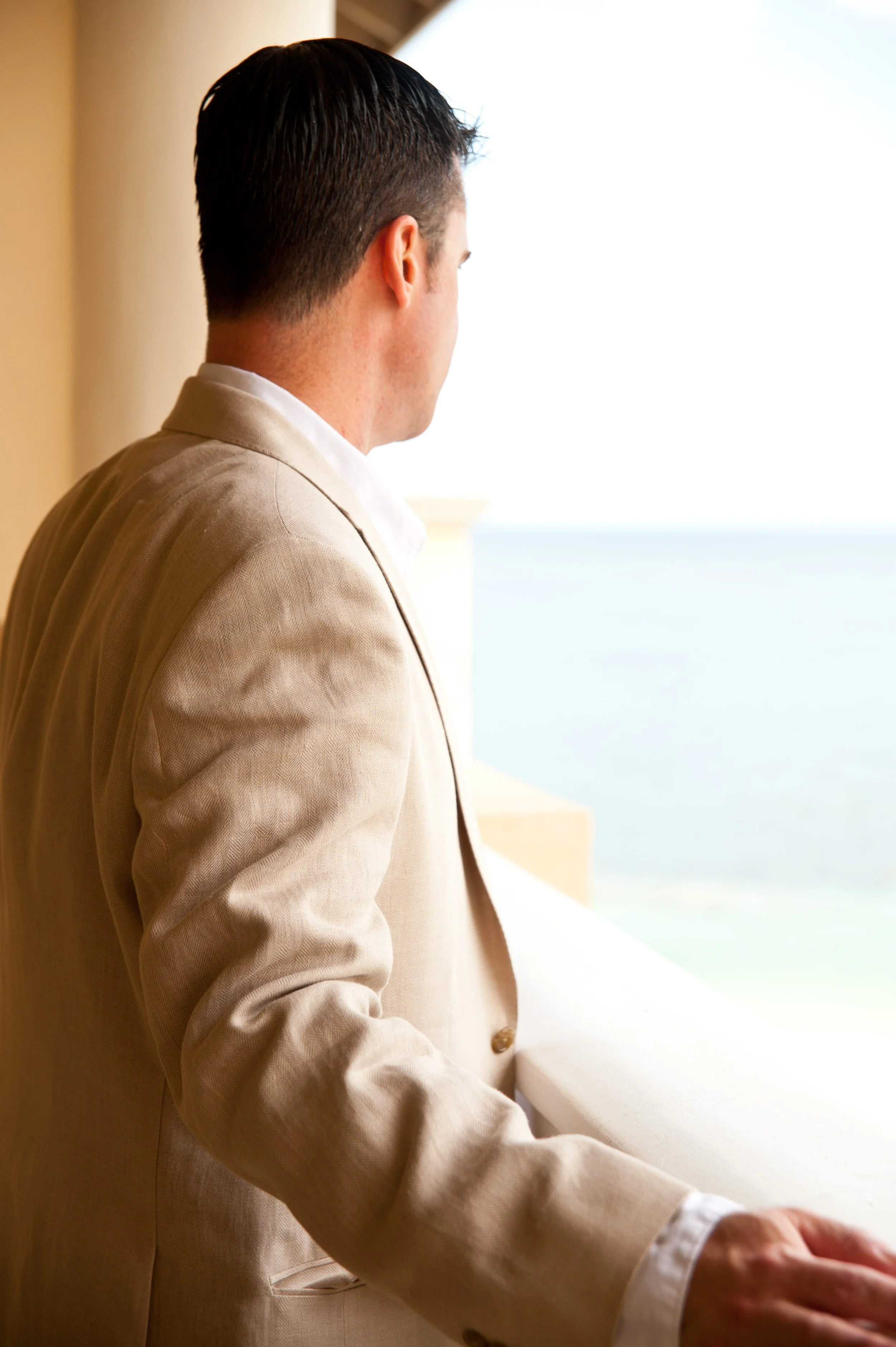 A man in a beige suit looking out a window at the ocean.