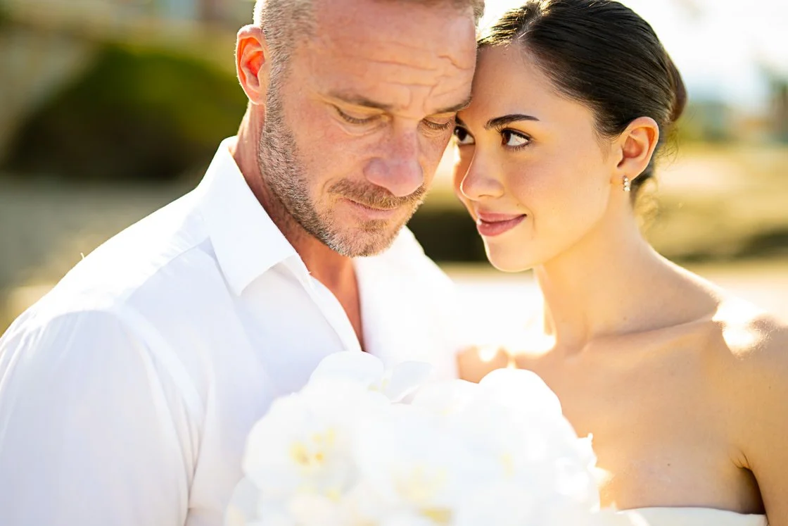 A couple, a man with a beard and woman with dark hair, are close together outdoors, holding a white wedding cake. They are dressed in white, and the background is blurred with warm sunlight.