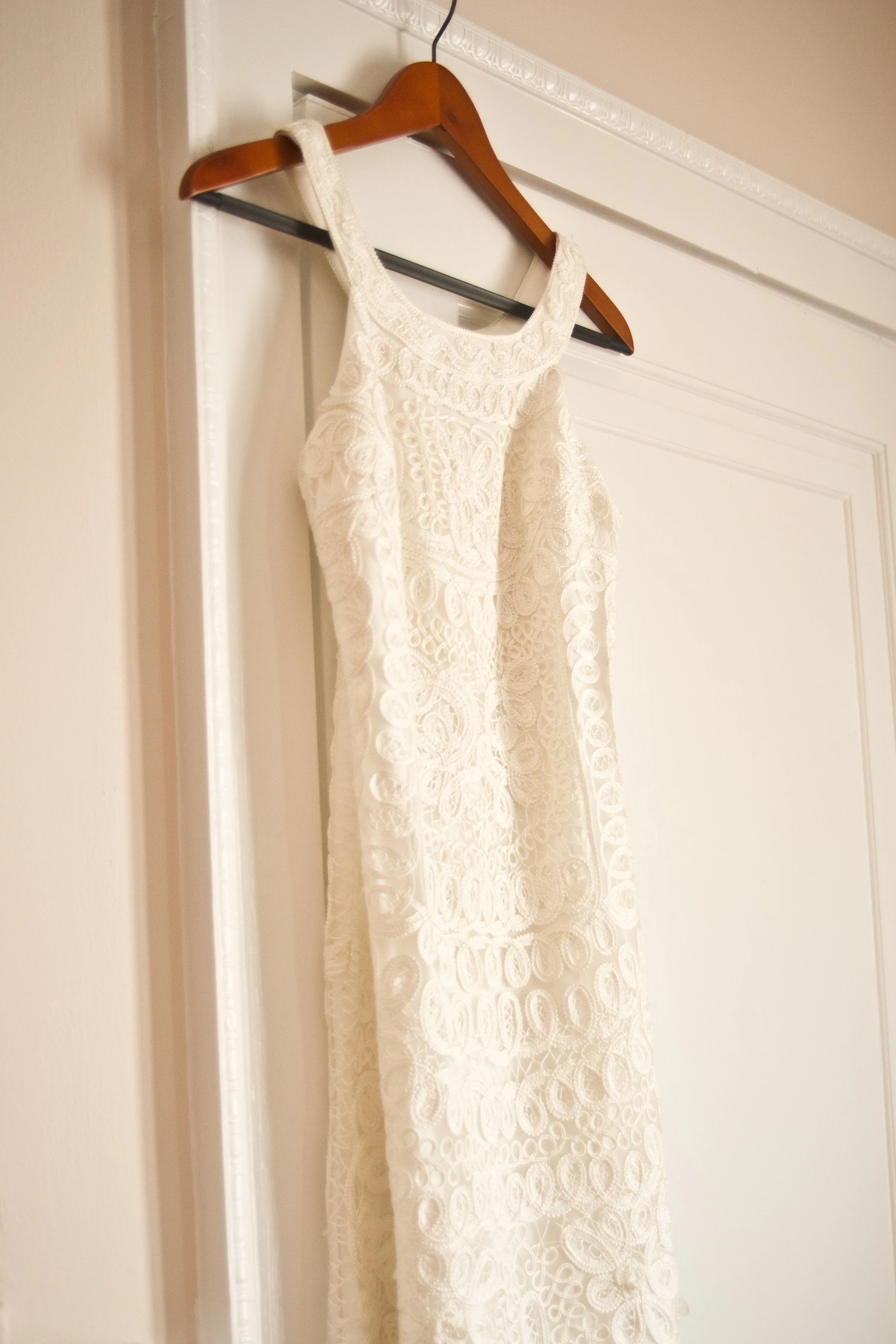 A white lace dress hanging on a wooden hanger on a metal bar inside a room with white walls and decorative crown molding.