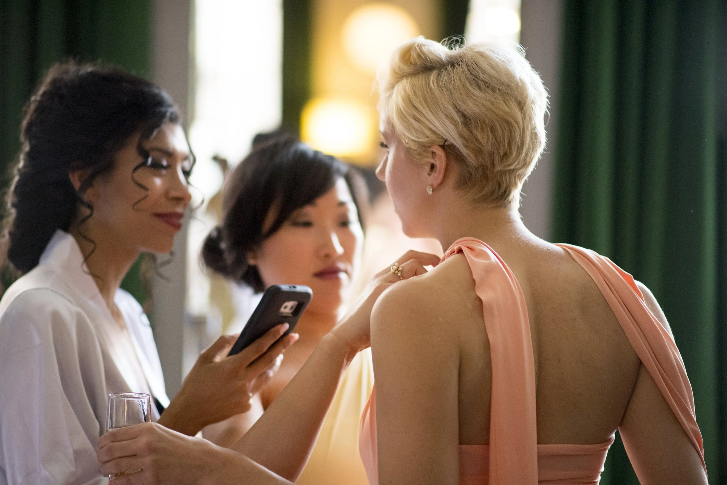 Three women are standing close together indoors. The woman on the left has dark curly hair and glasses, holding a phone, and is wearing a white top. The woman in the middle has dark straight hair, and the woman on the right has short blonde hair, ear