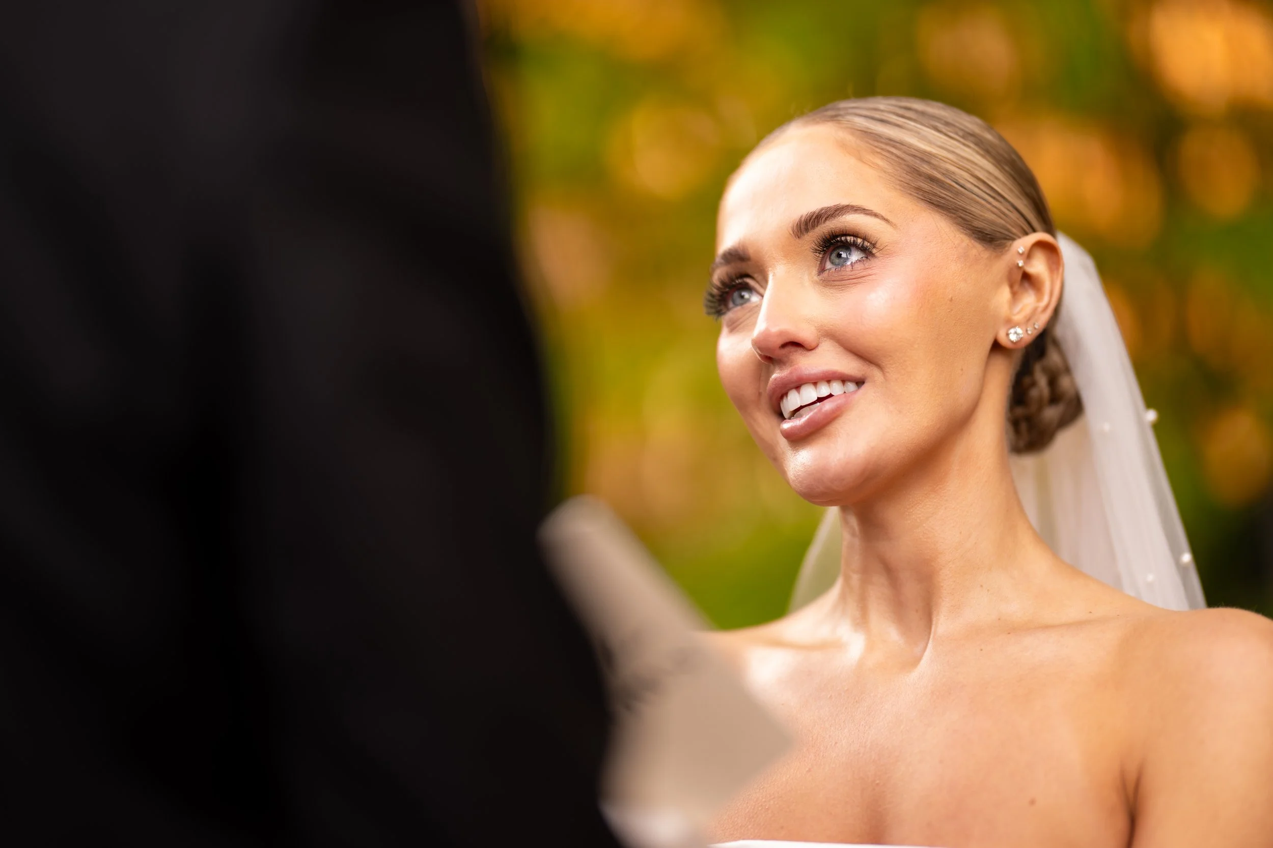 A woman with blue eyes, smiling, with braided hair and ear jewelry, looks at a person in black attire in an outdoor setting with autumn-colored blurred background.