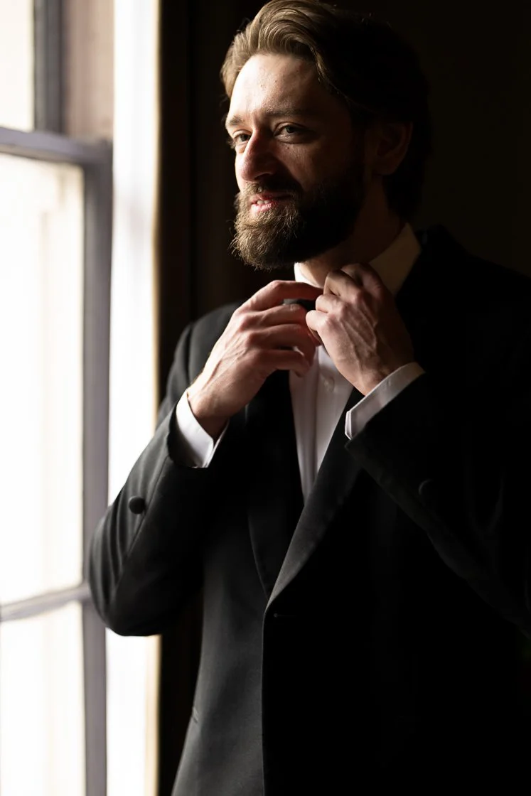 A man adjusting his bow tie in a dimly lit room by a window.