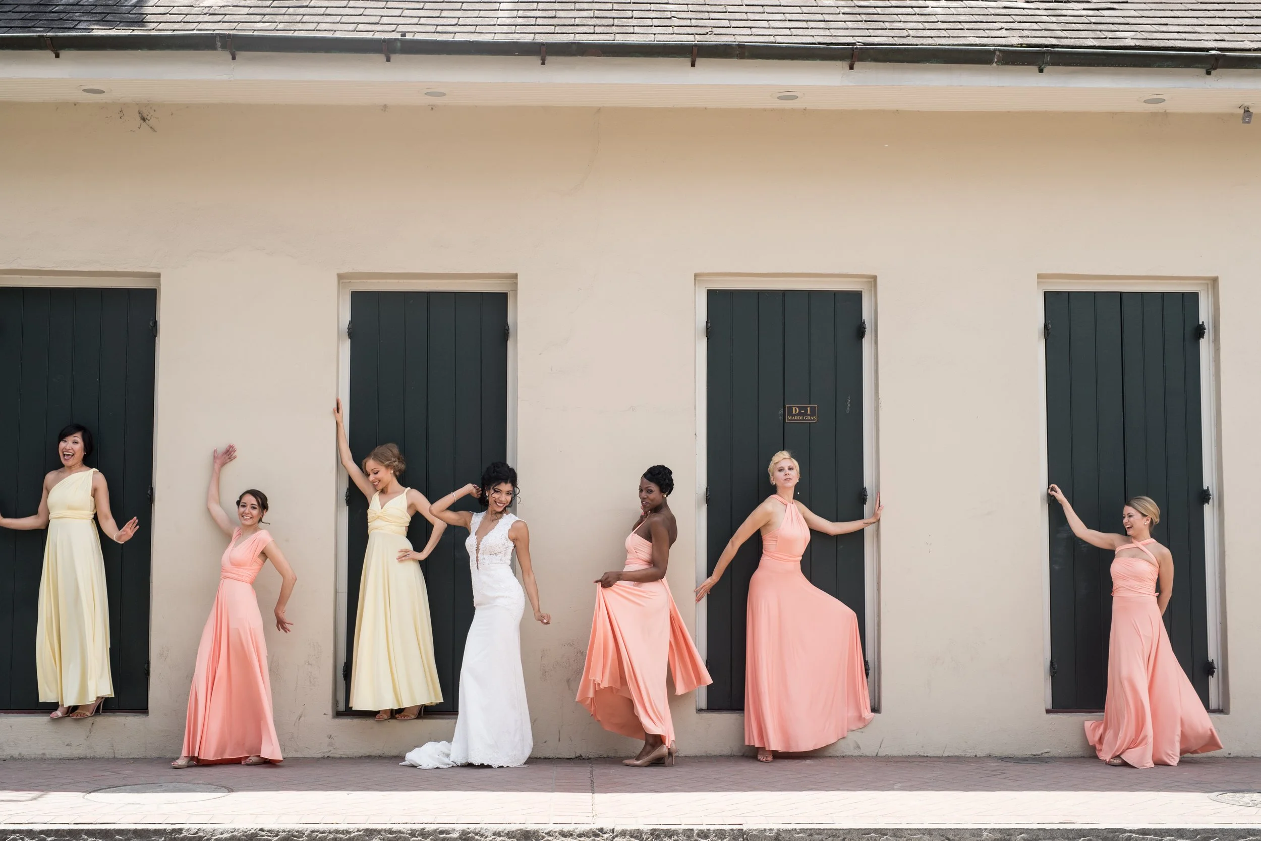 A group of women in colorful dresses posing in front of a beige wall with three black doors, celebrating a wedding.