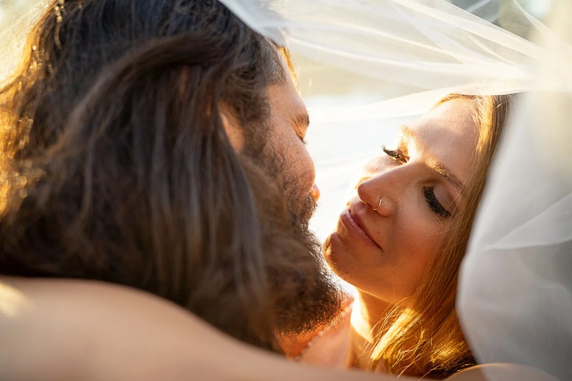 Close-up of a couple lying close together, with their faces inches apart, in warm sunlight. The woman has blonde hair, a nose ring, and closed eyes; the man has long dark hair, a beard, and closed eyes.