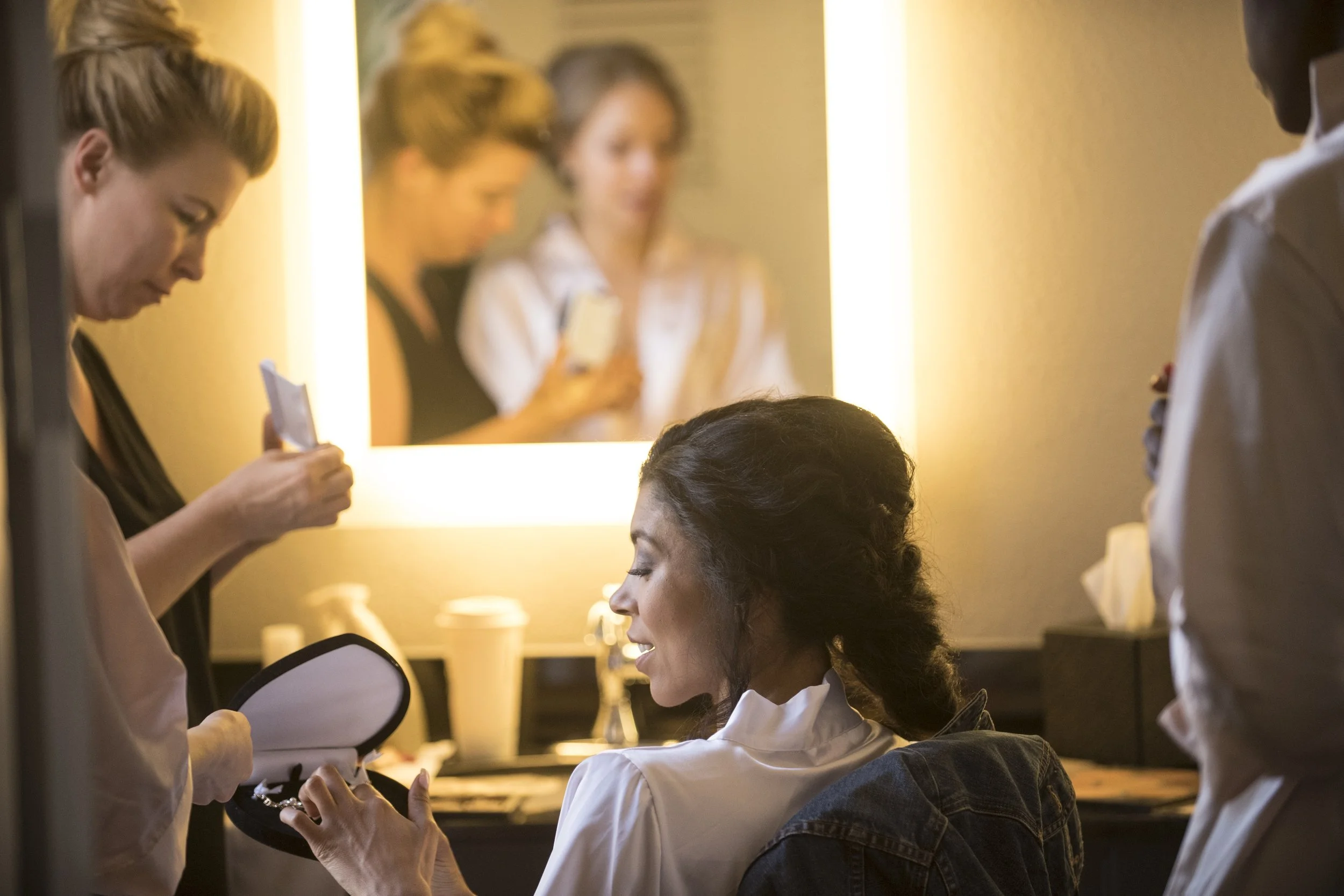 A woman helps another woman with her makeup in front of a mirror in a salon, surrounded by hairstylists and salon staff.