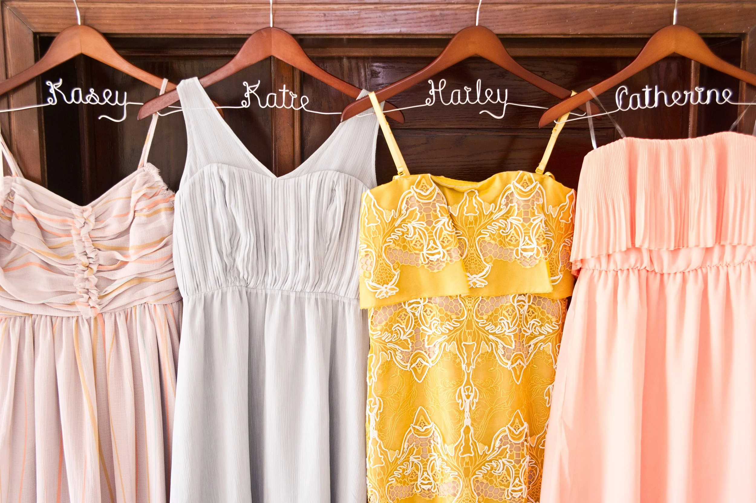 Clothing items hanging on a wooden rack, with wire hangers labeled with names, including Kasie, Katie, Harley, and Catherine. The clothes are dresses or blouses in pastel colors and patterns.