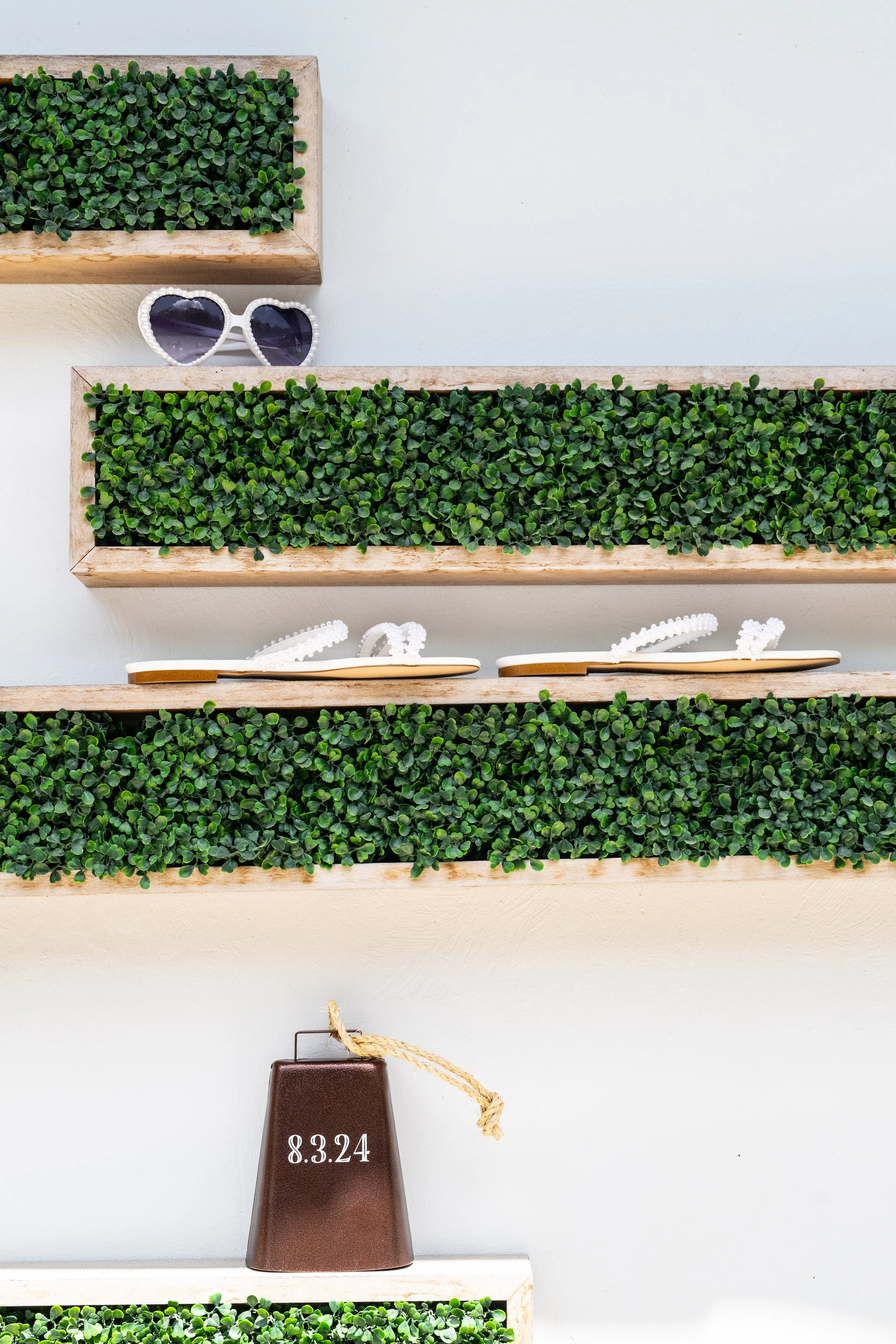 Wall shelves with faux greenery, white sunglasses, decorative element with the date 8.3.24, and a brown vase with a rope.