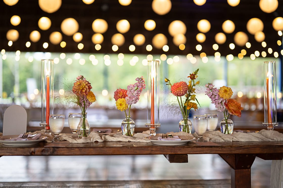 Wedding or event reception table decorated with colorful flowers in glass vases, tall candles in glass holders, and set with plates, glasses, and silverware, with string lights overhead.