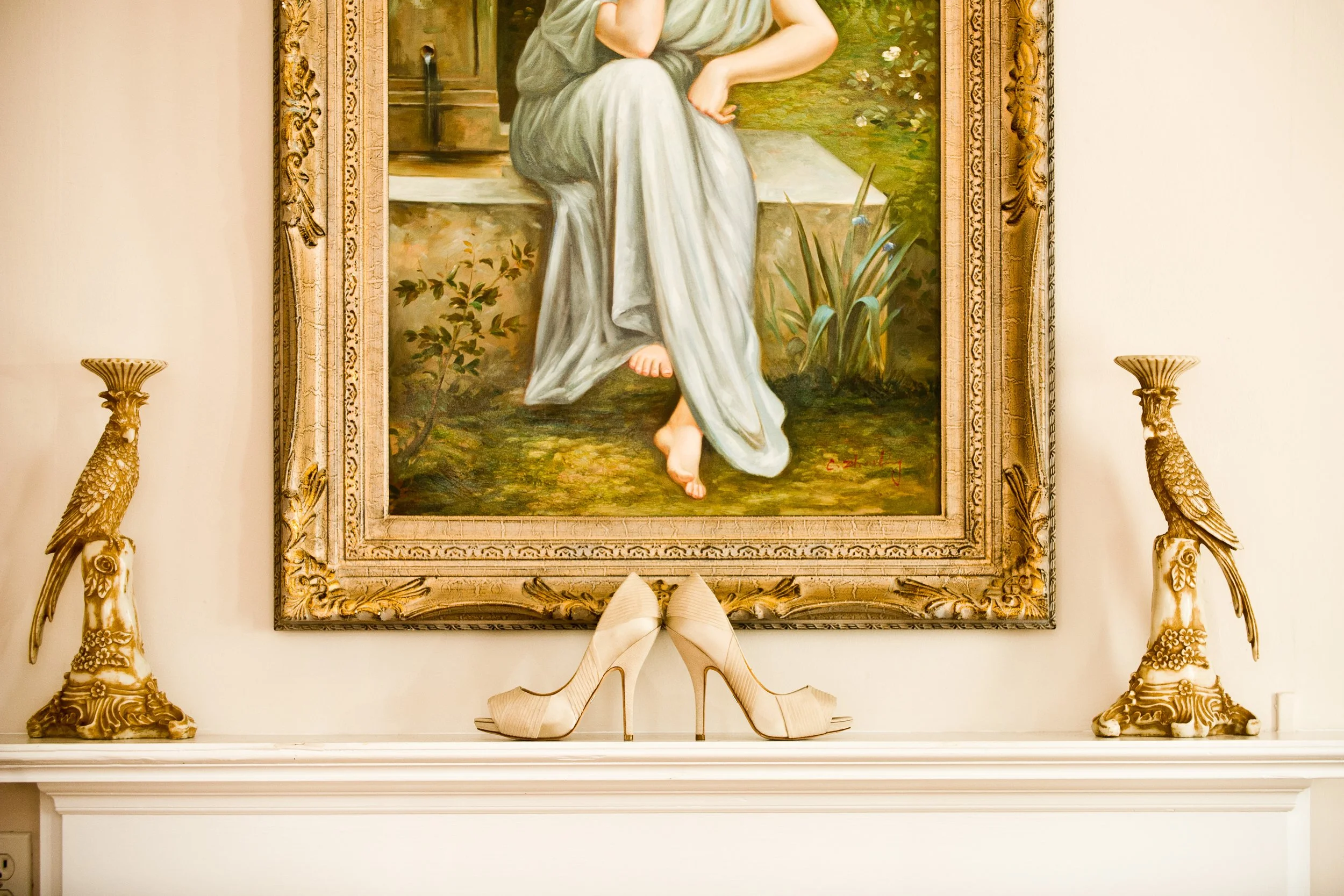 A framed painting of a woman sitting outdoors, with her hand on her chin and a relaxed pose, displayed on a white wall flanked by ornate peacock-shaped gold candle holders.
