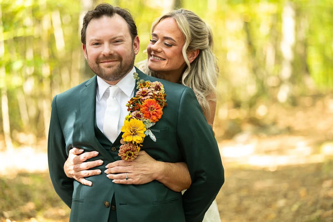 A couple in wedding attire outdoors, the woman embracing the man and both smiling, surrounded by trees and yellow foliage.
