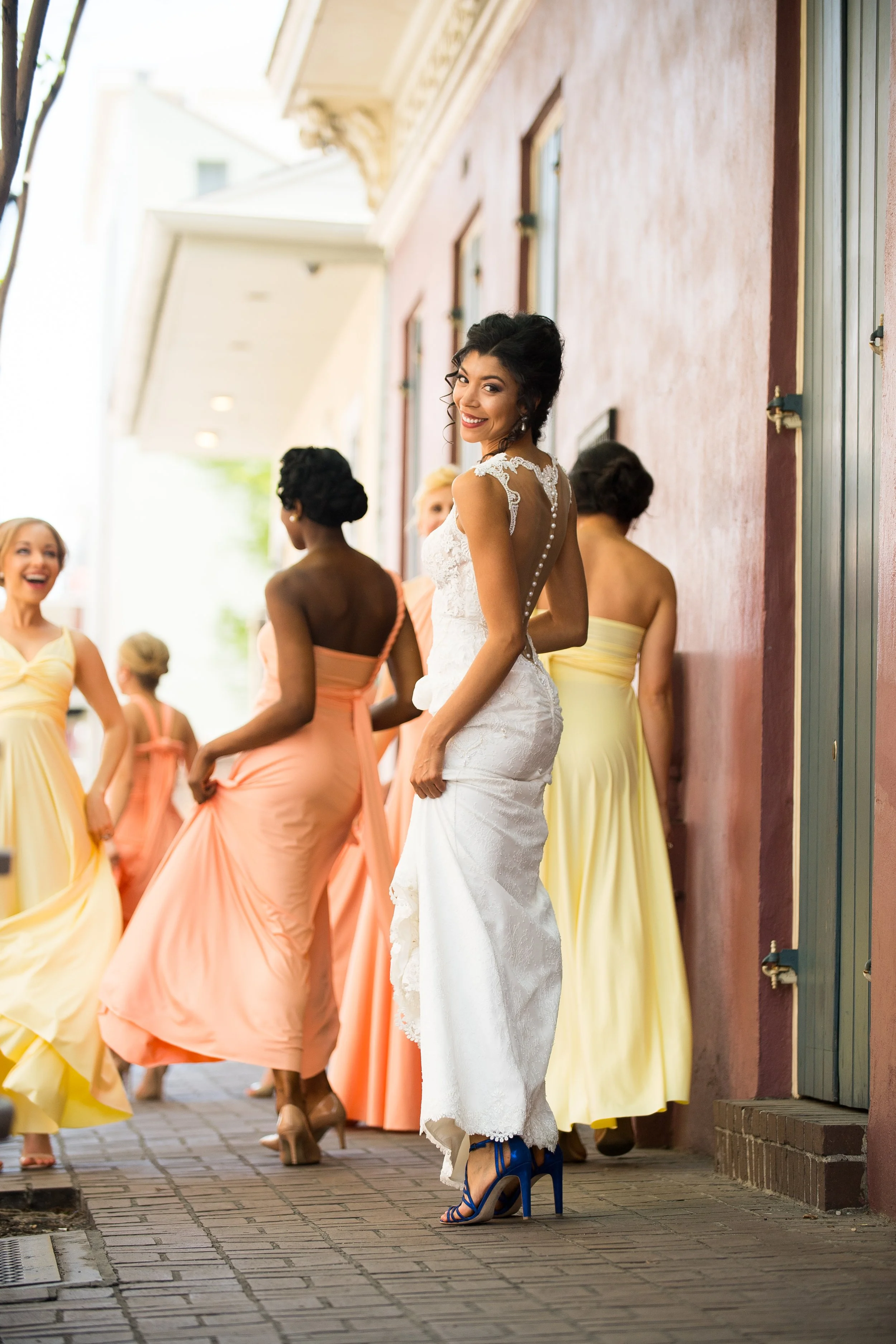 A bride in a white lace wedding gown with blue high heels and a lace-up ankle bracelet standing on a city sidewalk, surrounded by bridesmaids in pastel-colored dresses.
