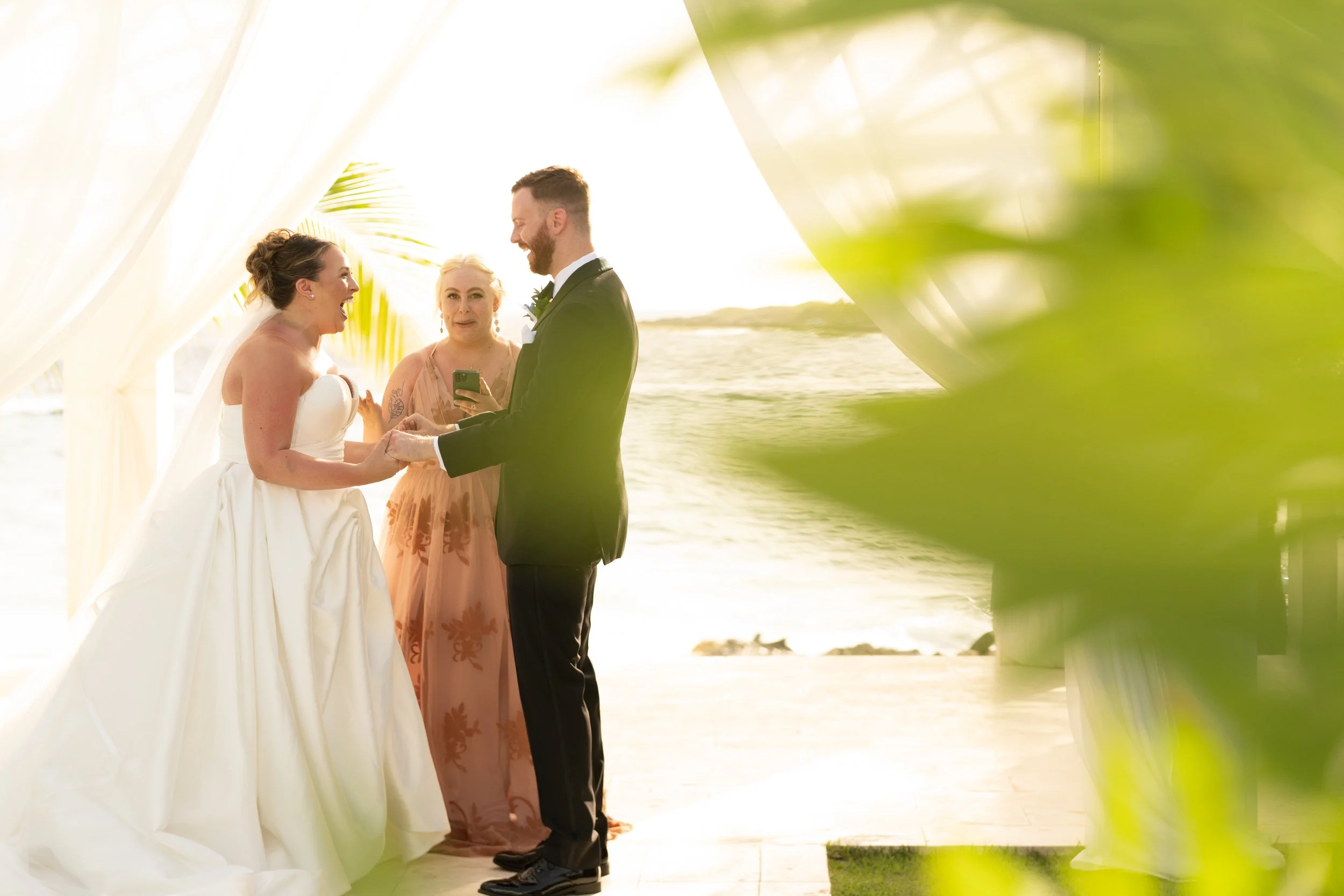 A wedding ceremony takes place outdoors on a beach with the ocean in the background, featuring a bride and groom holding hands, smiling, and a woman officiant in the middle. The scene is framed with greenery.