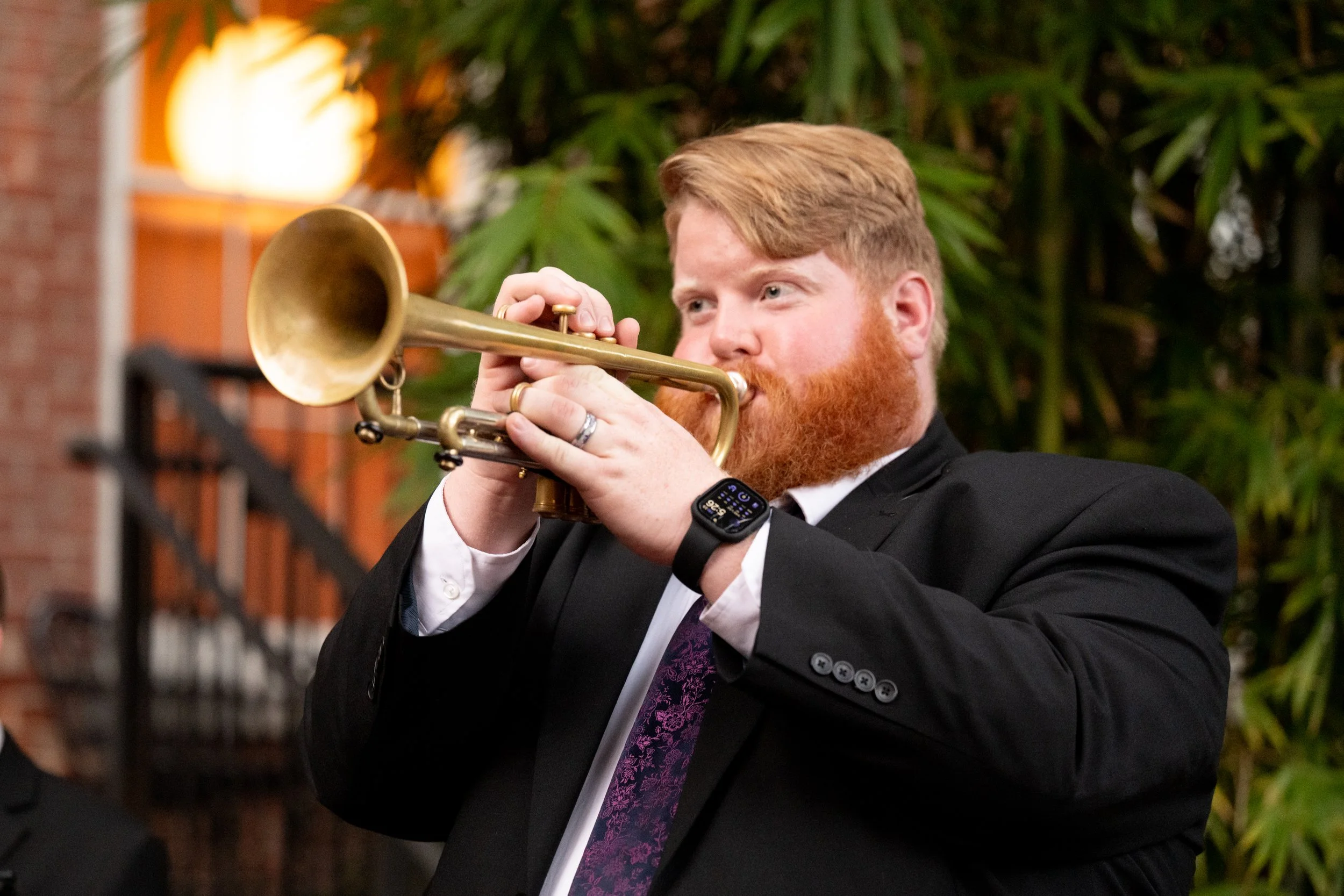 A man with red hair and beard wearing a black suit and purple tie playing a brass trumpet outdoors at night.