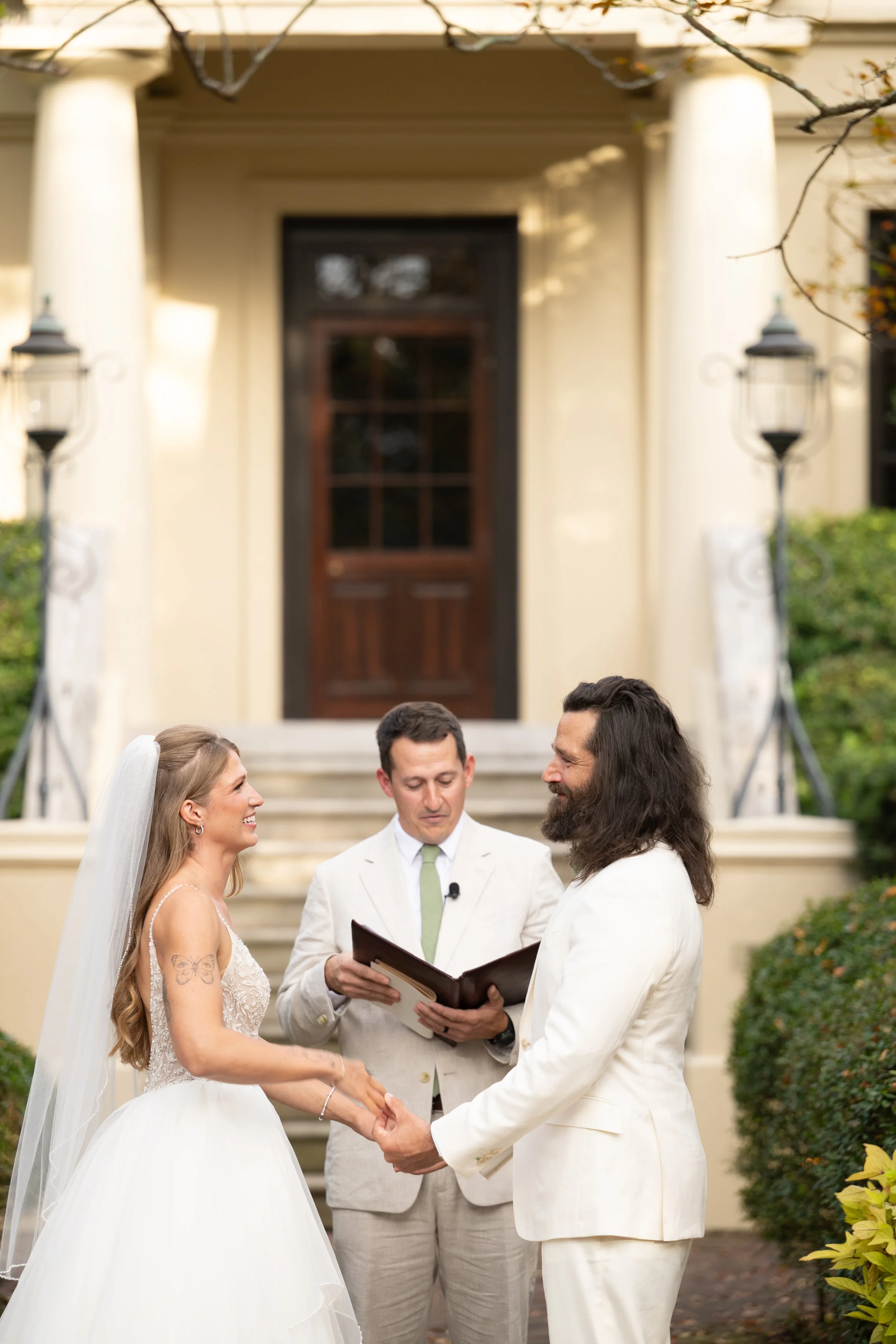 A wedding ceremony outdoors with a bride and groom holding hands and facing each other, couples exchanging vows, officiant reading from a book, and an elegant building with stairs and greenery in the background.