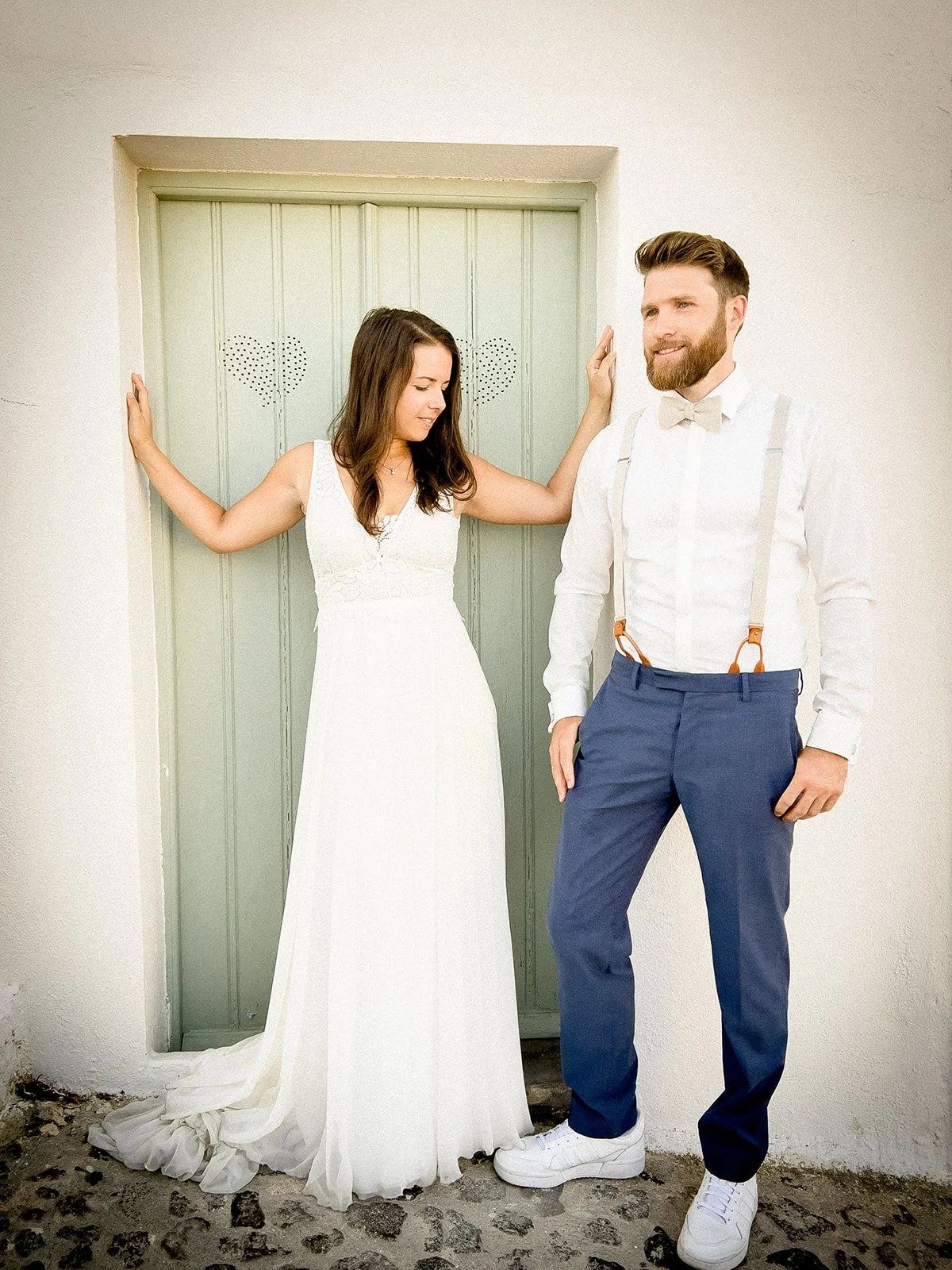 A bride and groom pose in front of a green door decorated with two hearts. The bride is dressed in a white wedding gown, and the groom is wearing a white shirt with suspenders, blue pants, and white sneakers.