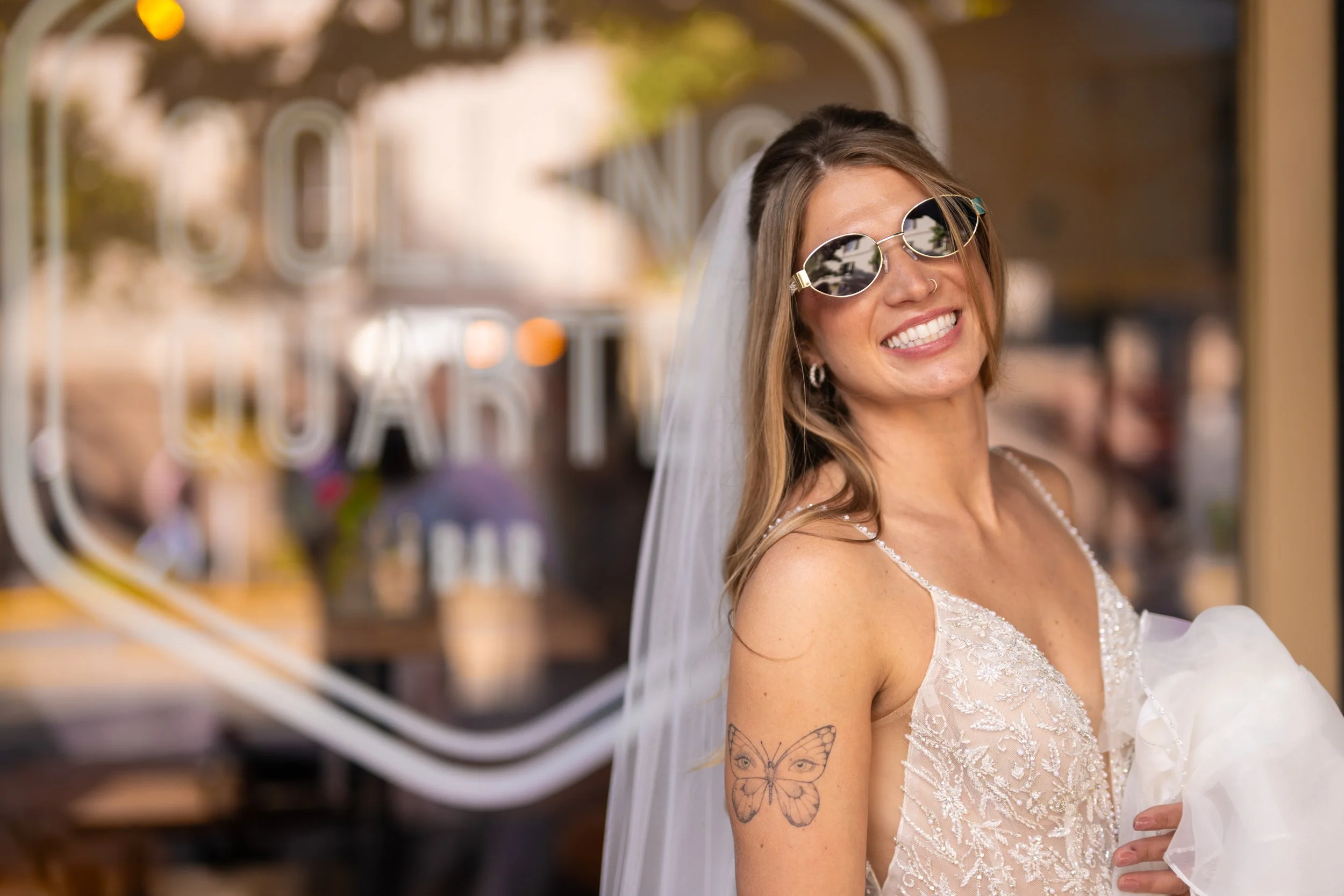 Close-up of a smiling woman wearing sunglasses and a white lace wedding dress, with visible butterfly tattoos on her arm, standing in front of store window with blurred background.