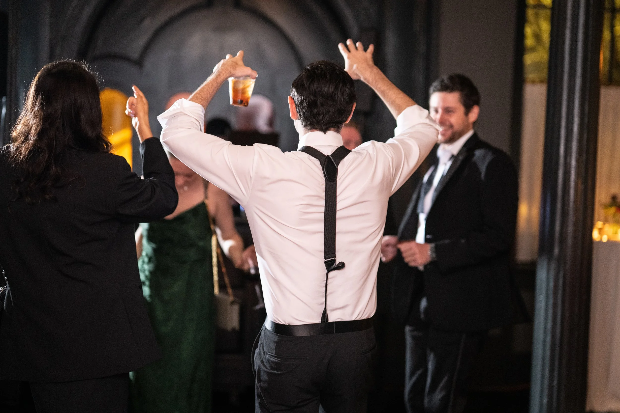 People dancing at a social event or celebration, with a man in the center raising his arms and holding a drink, while others smile and enjoy the moment.