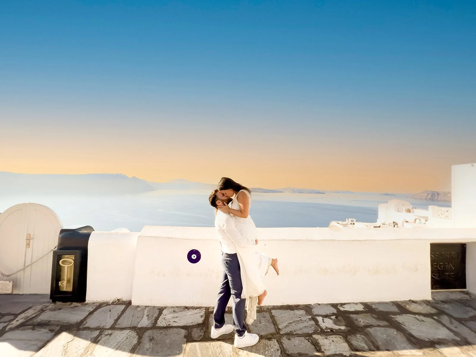 A couple kissing on a rooftop overlooking the ocean during sunset, with white buildings and a stone pathway in the foreground.