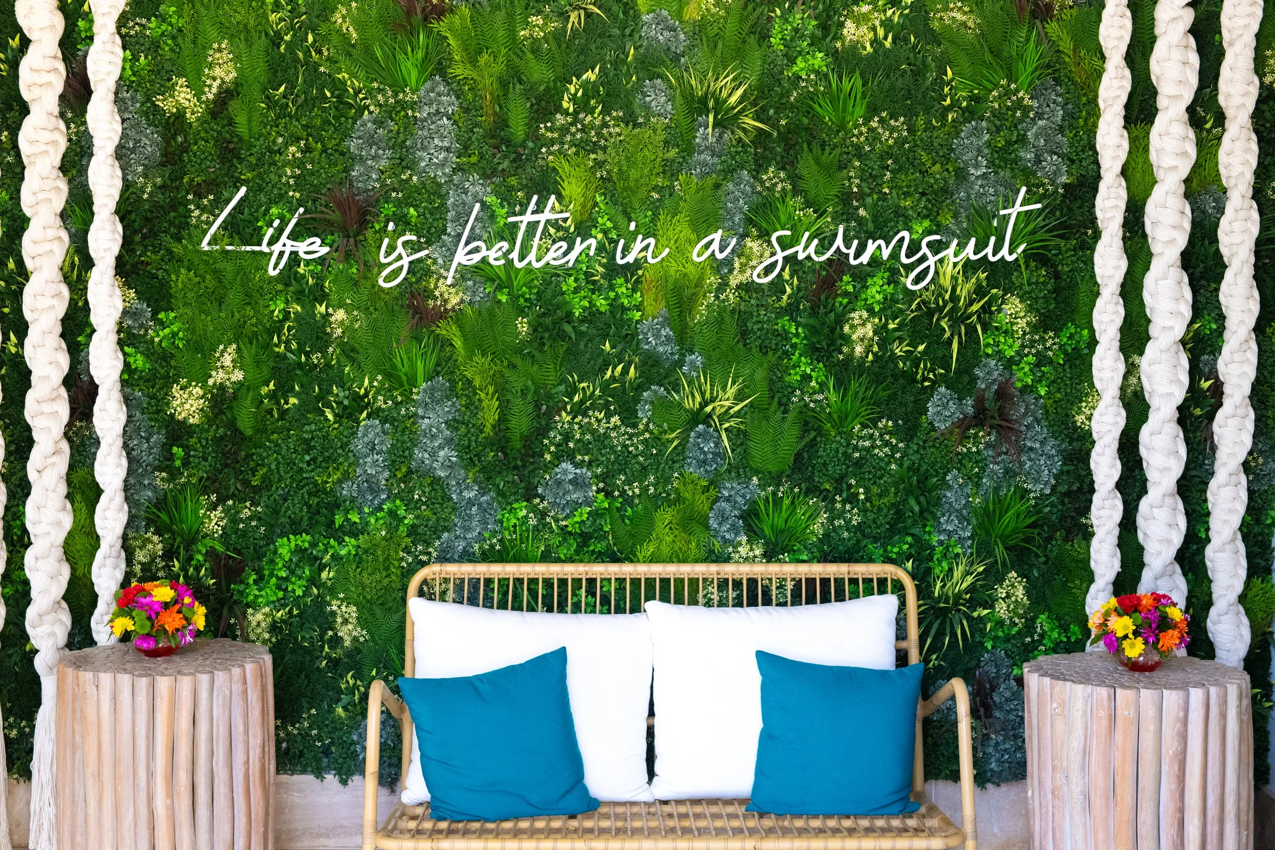 A green wall with plants and white cursive text that reads "Life is better in a sunmout". There are two small tables with flowers on either side of a white sofa with blue cushions, and white hanging ropes on either side of the wall.