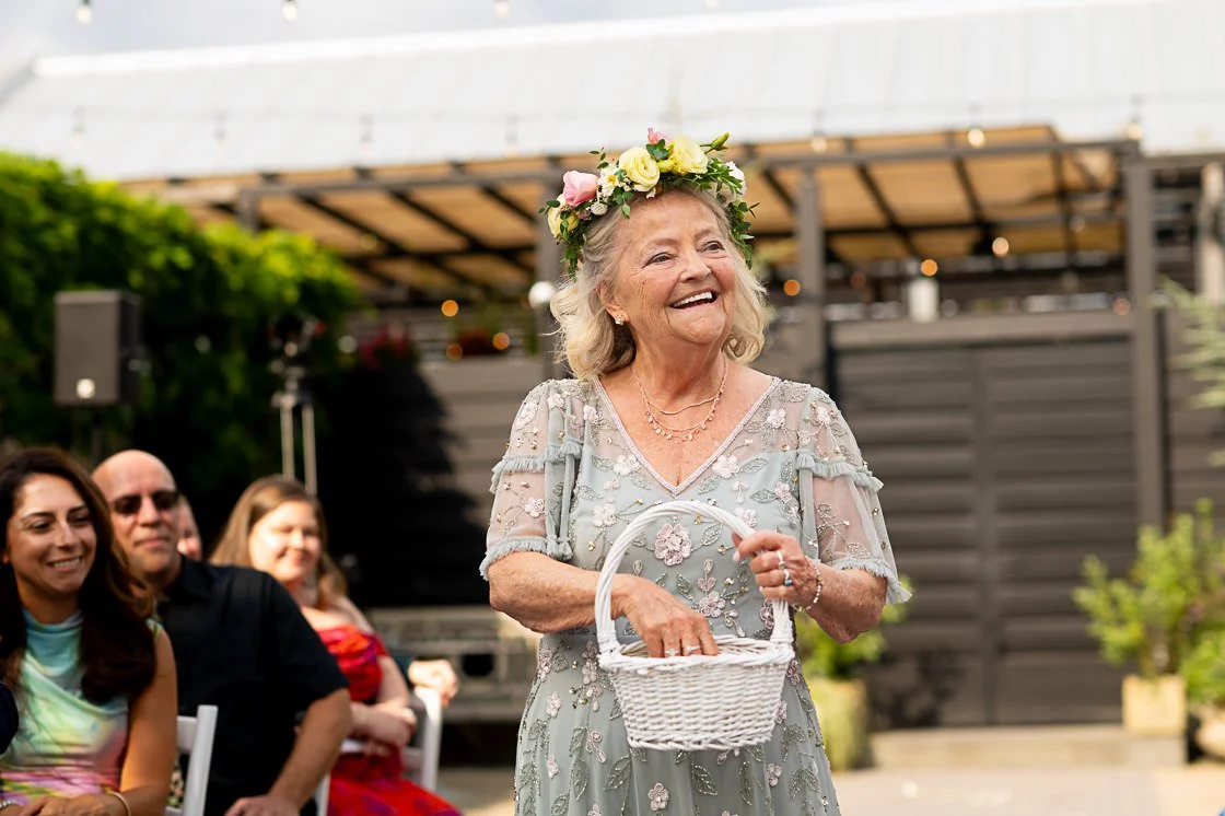 An elderly woman with a floral crown and a light gray, embroidered dress holding a white basket, smiling at an outdoor gathering.