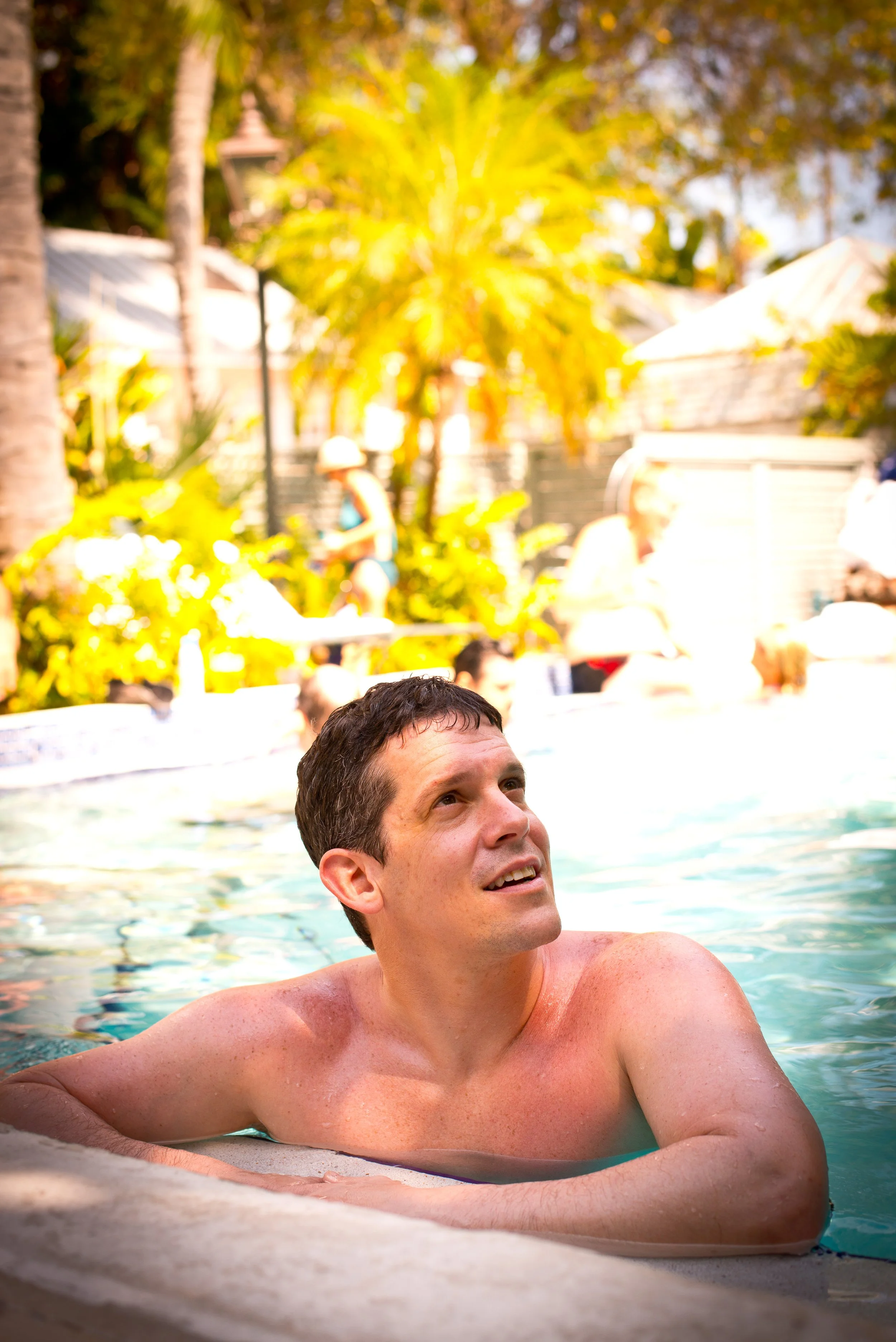 A man with short dark hair is relaxing in a swimming pool with clear water, leaning on the pool's edge with his arms extended. He is smiling slightly and looking upwards. In the background, there are lush trees, palm trees, and a sunny yard with peop