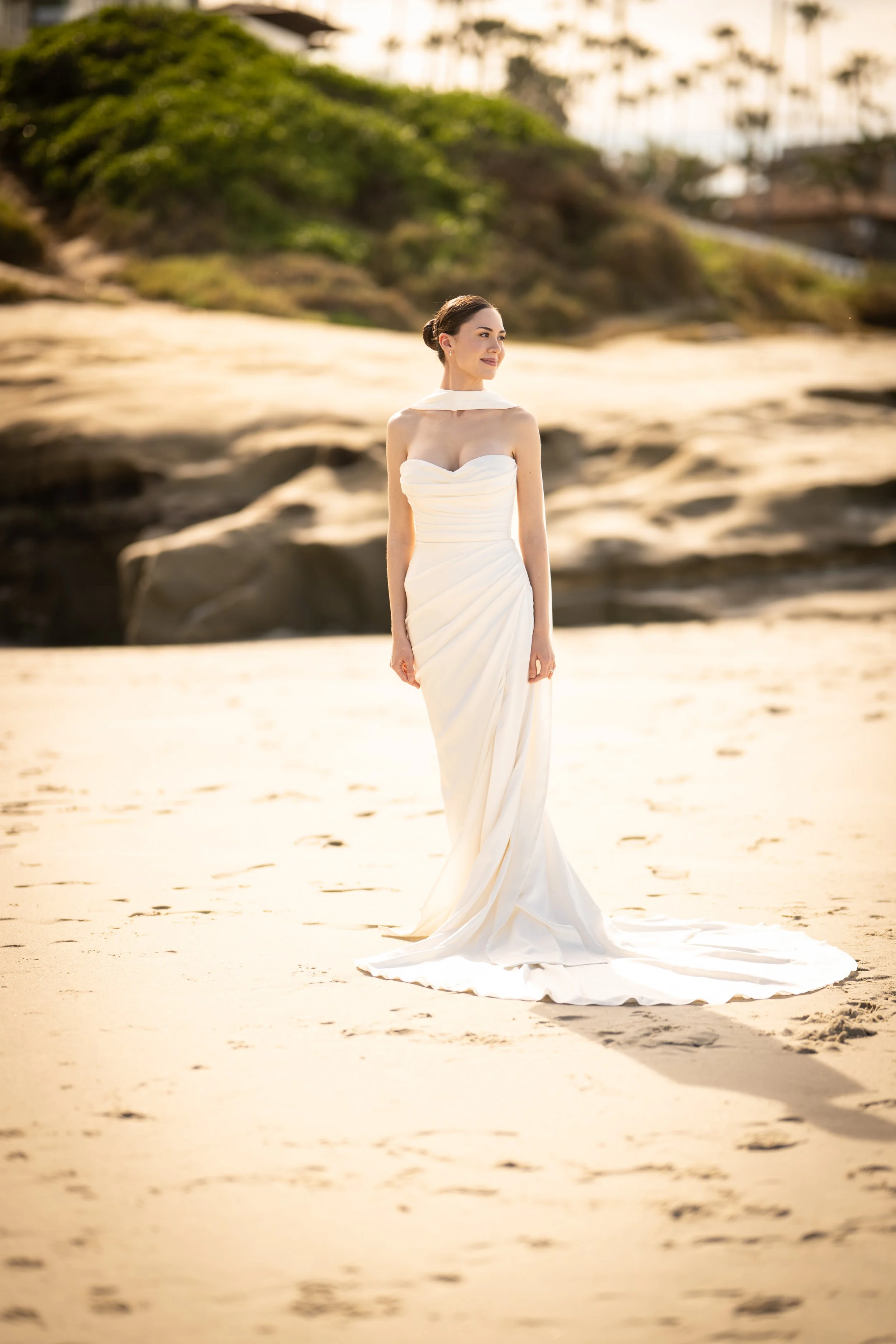 A woman in a white wedding dress standing on a sandy beach, with greenery and buildings in the background.