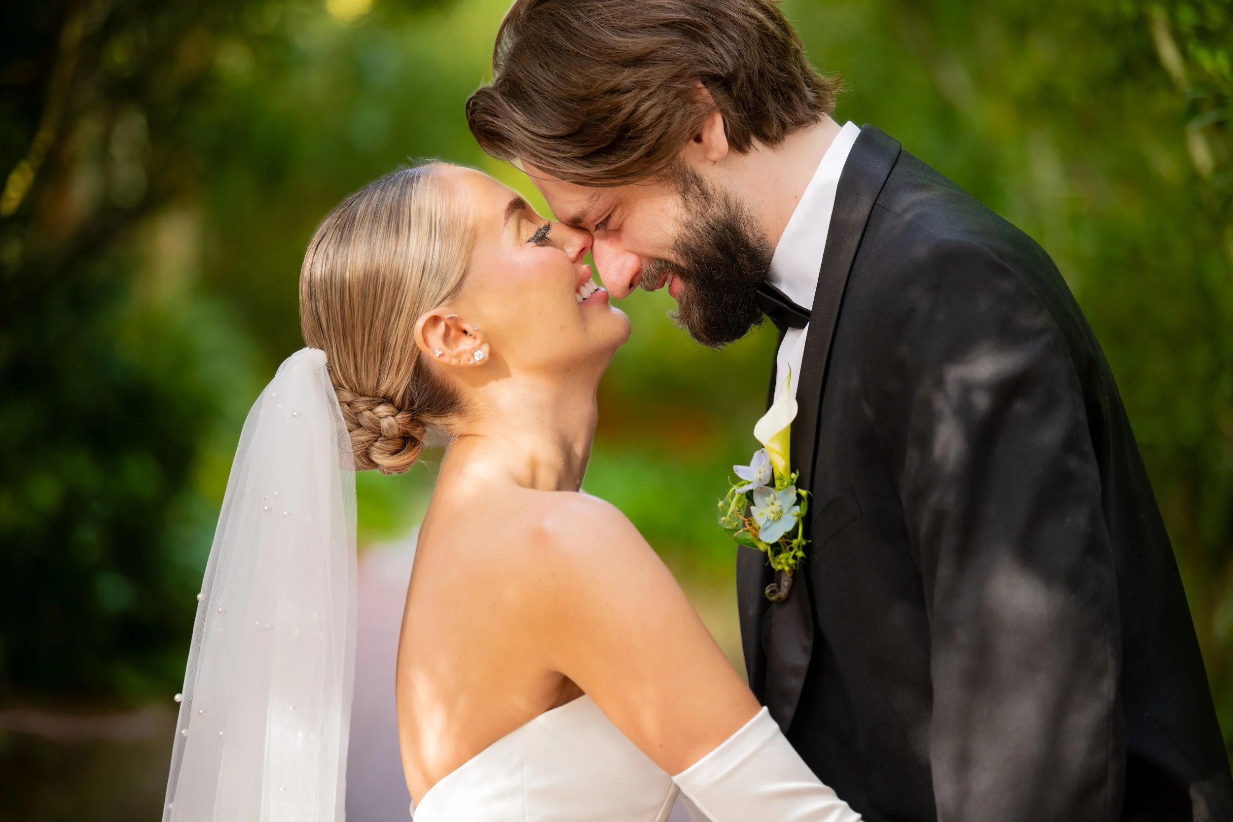A bride and groom in formal wedding attire touching foreheads and smiling at each other outdoors amid green trees.