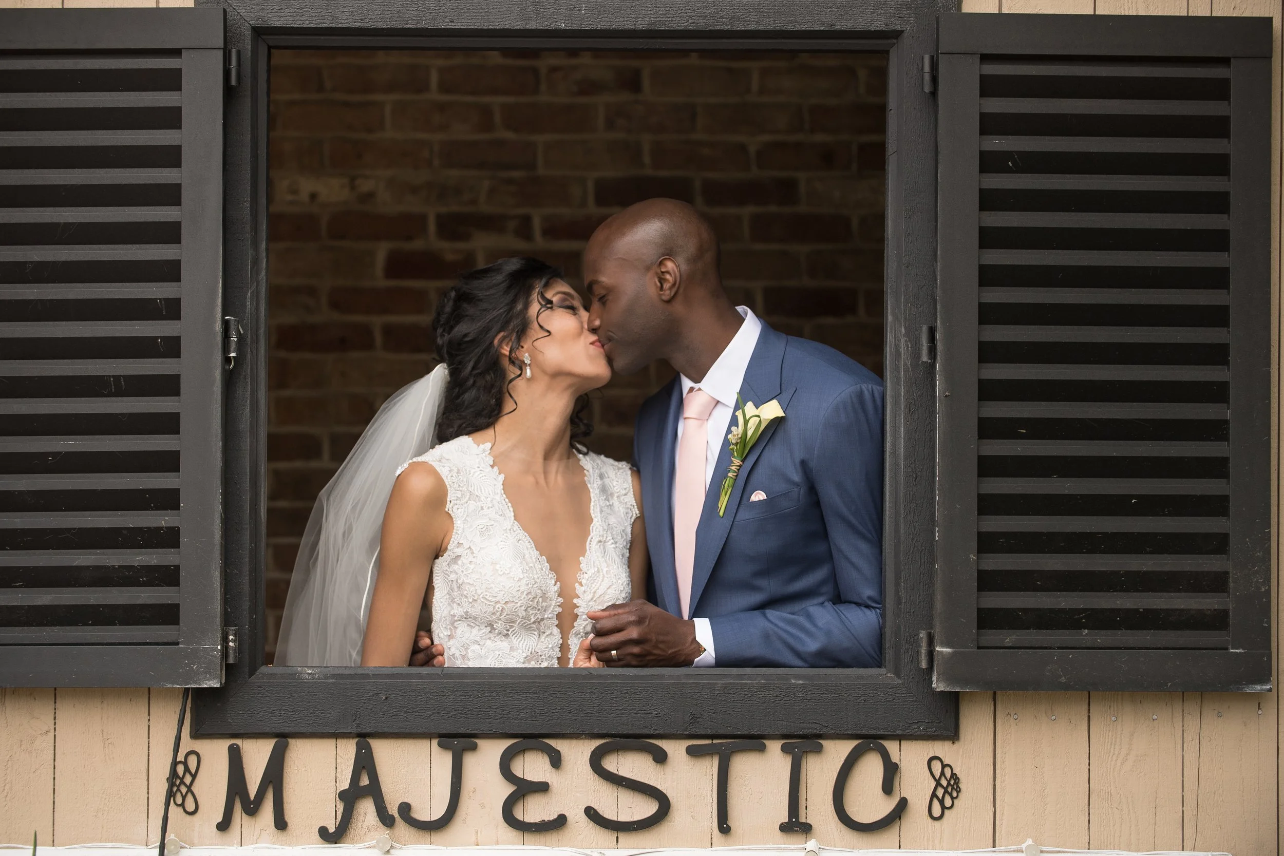 A couple in wedding attire sharing a kiss through a window. The bride is wearing a lace wedding dress with a veil, and the groom is wearing a blue suit with a pink tie, holding her hand.